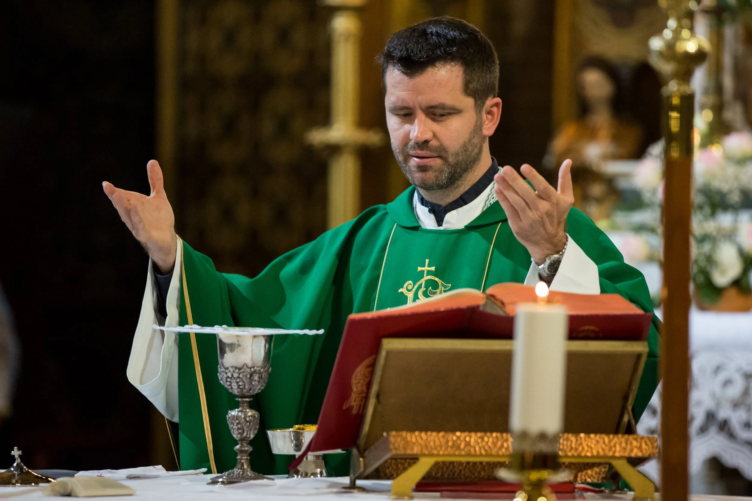 A priest dressed in green liturgical vestments stands at an altar with arms raised during a religious ceremony, with ornate decorations and religious items visible in the background.