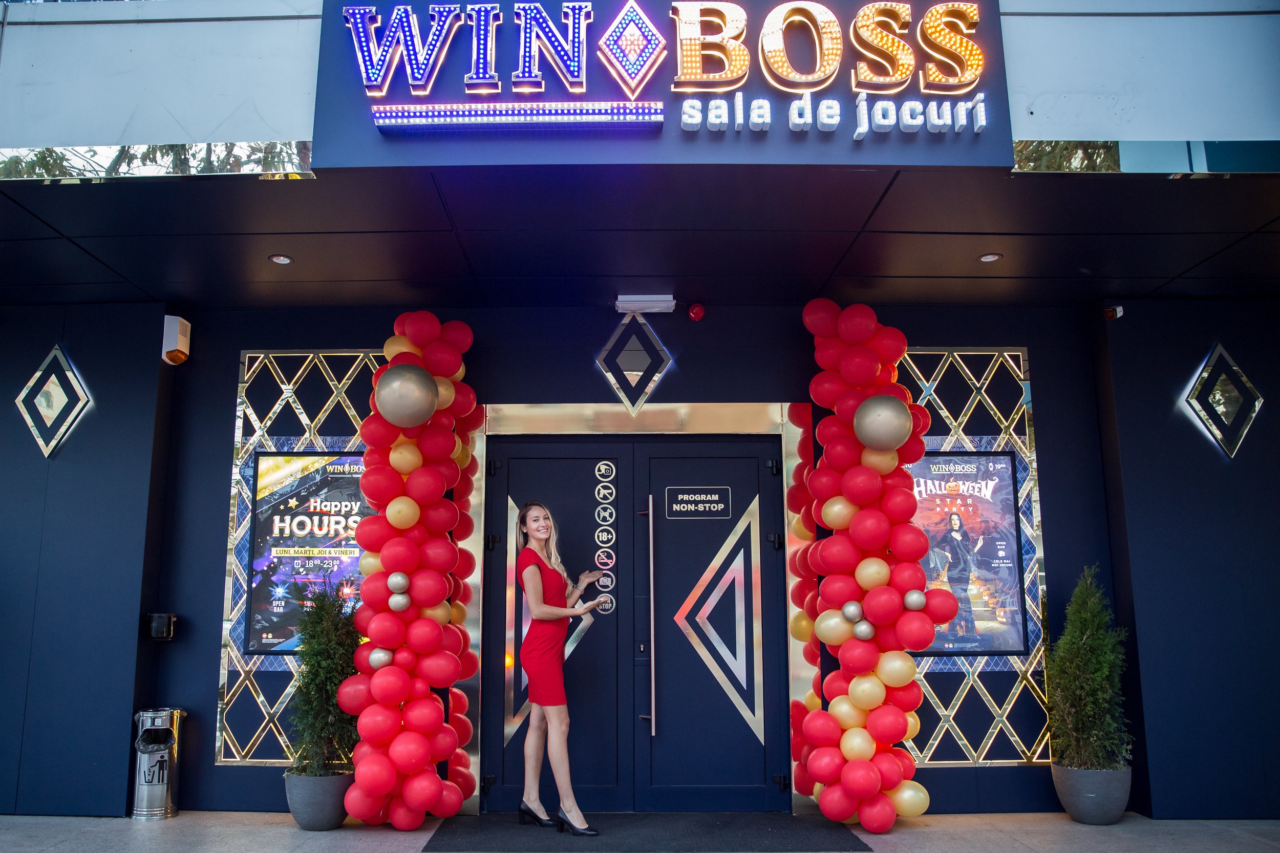 The entrance of a gaming room named WIN BOSS with a festive decoration. There are two columns of red, gold, and silver balloons framing the entrance. A woman dressed in a red dress and black high heels stands in front of the door, smiling and pointin