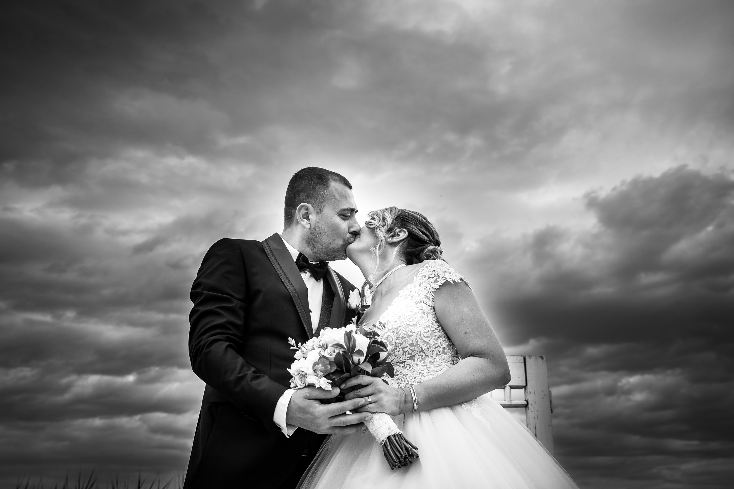 Black and white photo of a bride and groom kissing outdoors under a cloudy sky, holding a bouquet of flowers.