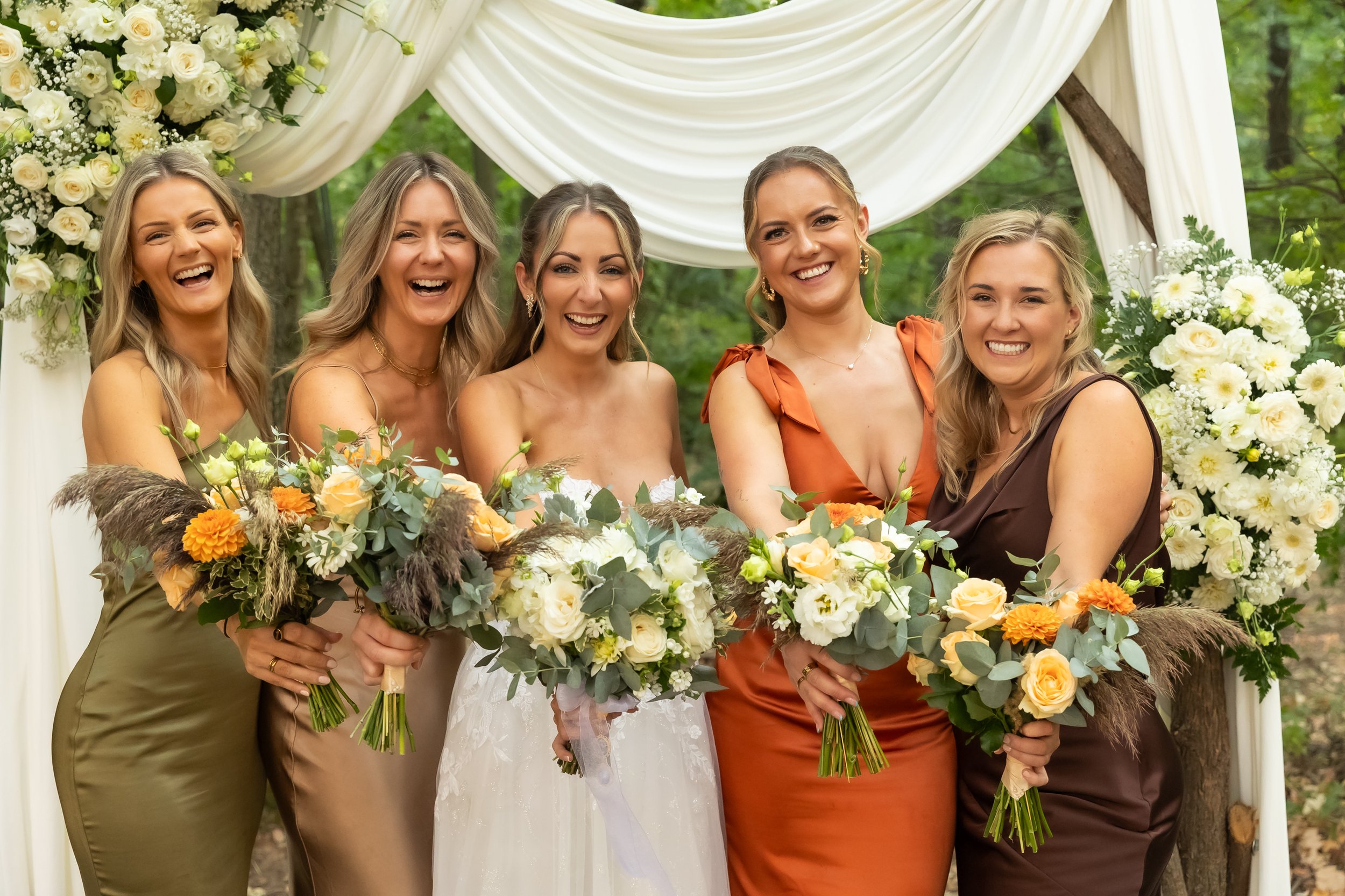A group of five women smiling and holding bouquets of flowers at an outdoor wedding ceremony. They are standing in front of a decorated arch with white drapes and floral arrangements, surrounded by trees.