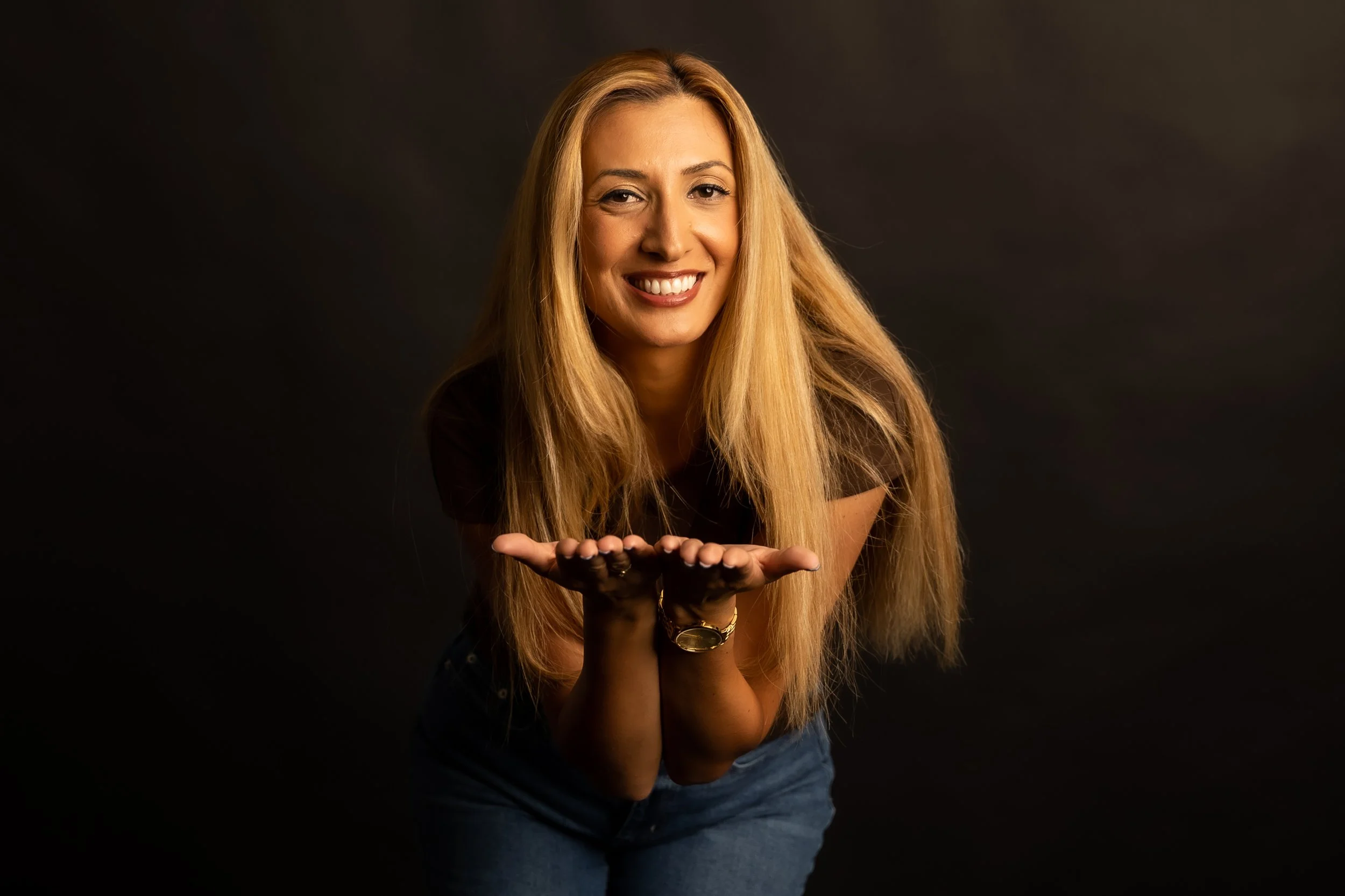 A woman with long blonde hair, smiling, leaning forward with her hands together, wearing a black shirt and blue jeans against a dark background.