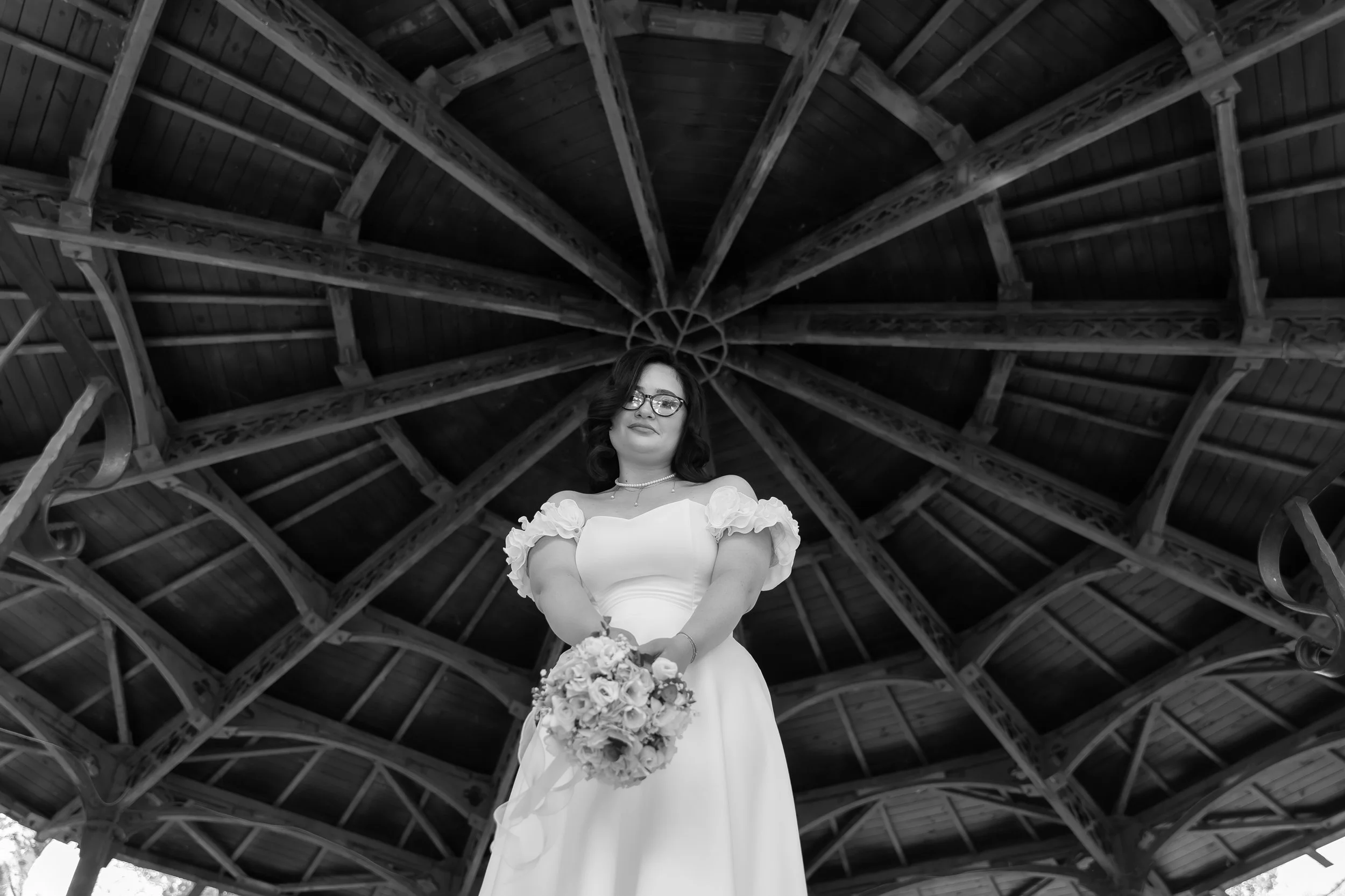 Black and white photo of a woman in a wedding dress holding a bouquet, standing inside a large wooden pavilion with intricate rafters overhead.