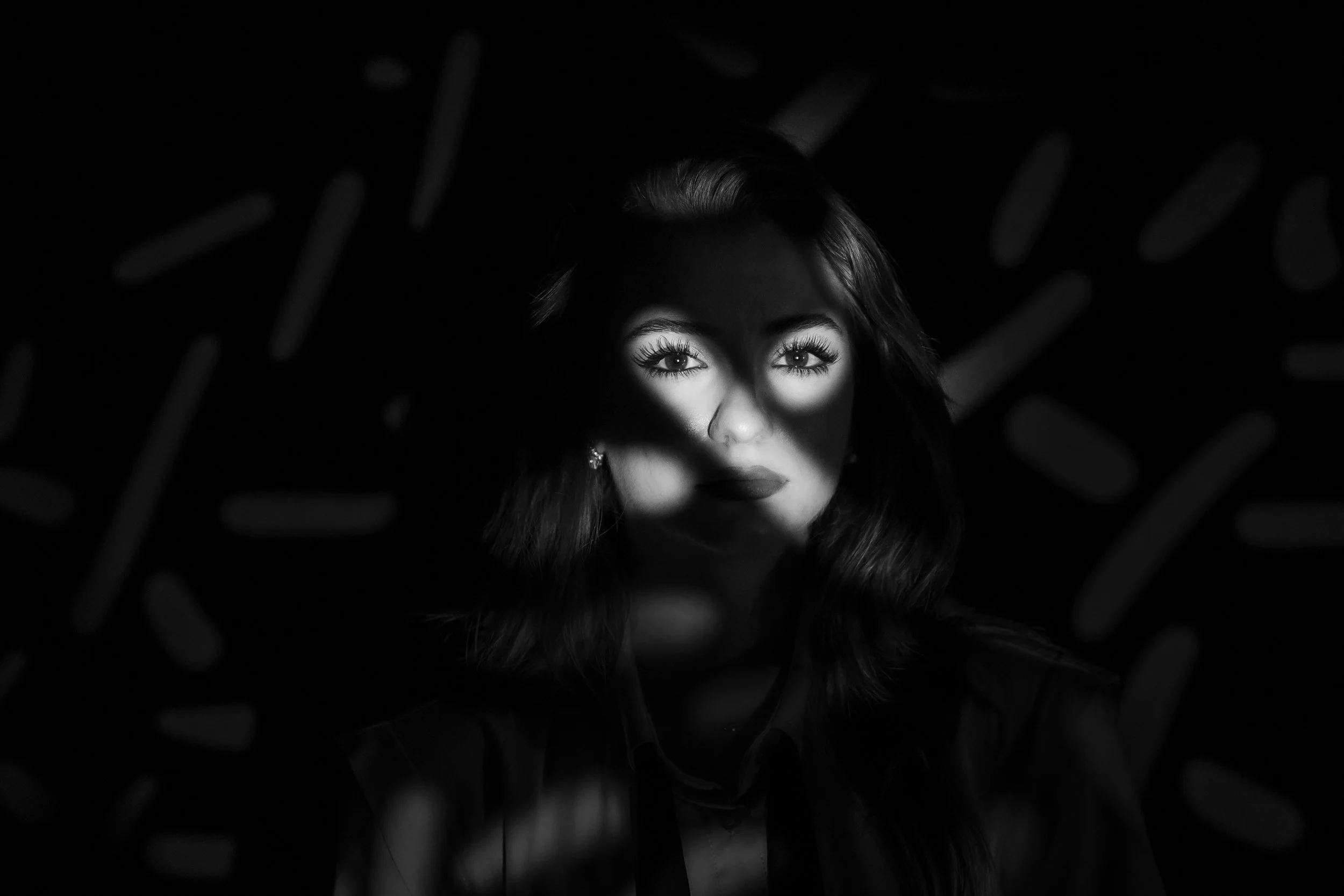 Black and white photo of a woman with long hair, looking directly at the camera, with light illuminating her eyes and face through a pattern of shadow and light.