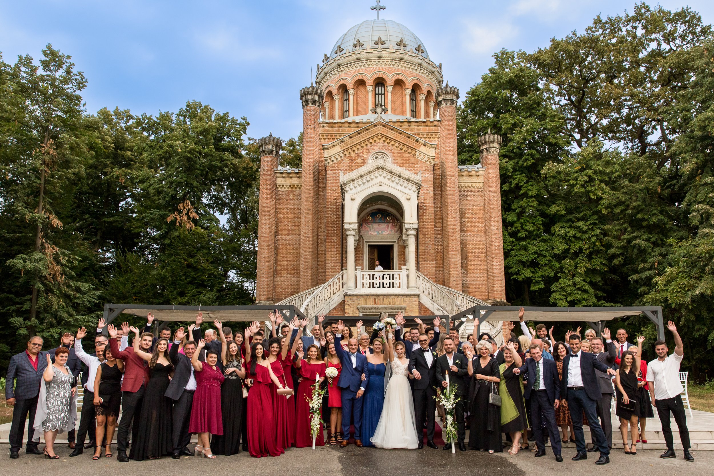 A large group of wedding guests and the bride and groom posing for a photo in front of a historic brick church with a domed steeple, surrounded by green trees.