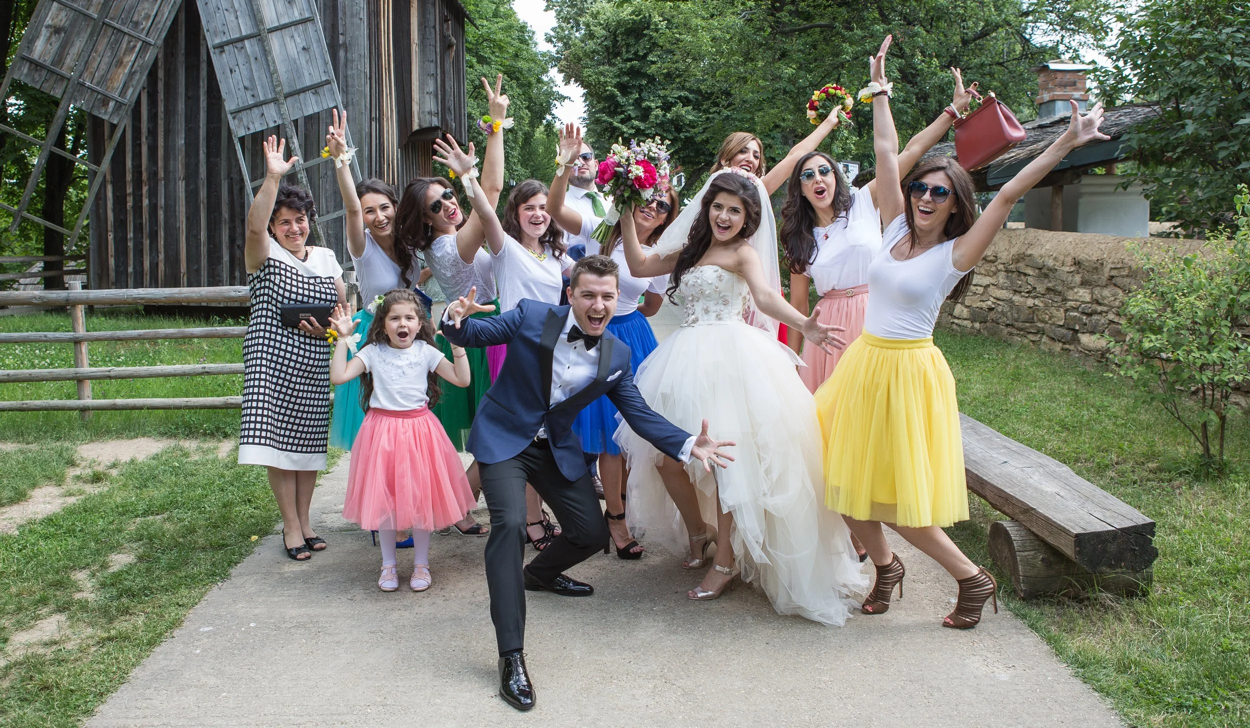 A group of people celebrating a wedding outdoors, with a bride in a white wedding dress and a groom in a tuxedo, surrounded by friends and family wearing colorful skirts and dresses, making joyful gestures and poses.