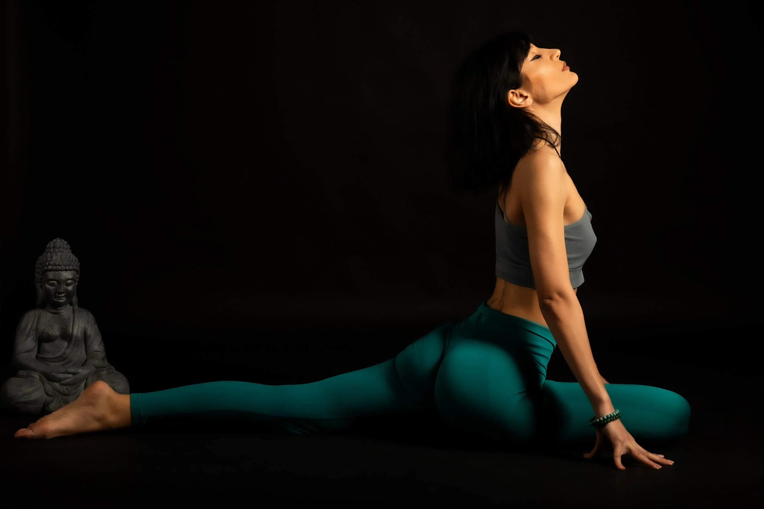 A woman practicing yoga in a cobra pose wearing teal leggings and a gray sports bra, with a Buddha statue on the left side of the black background.
