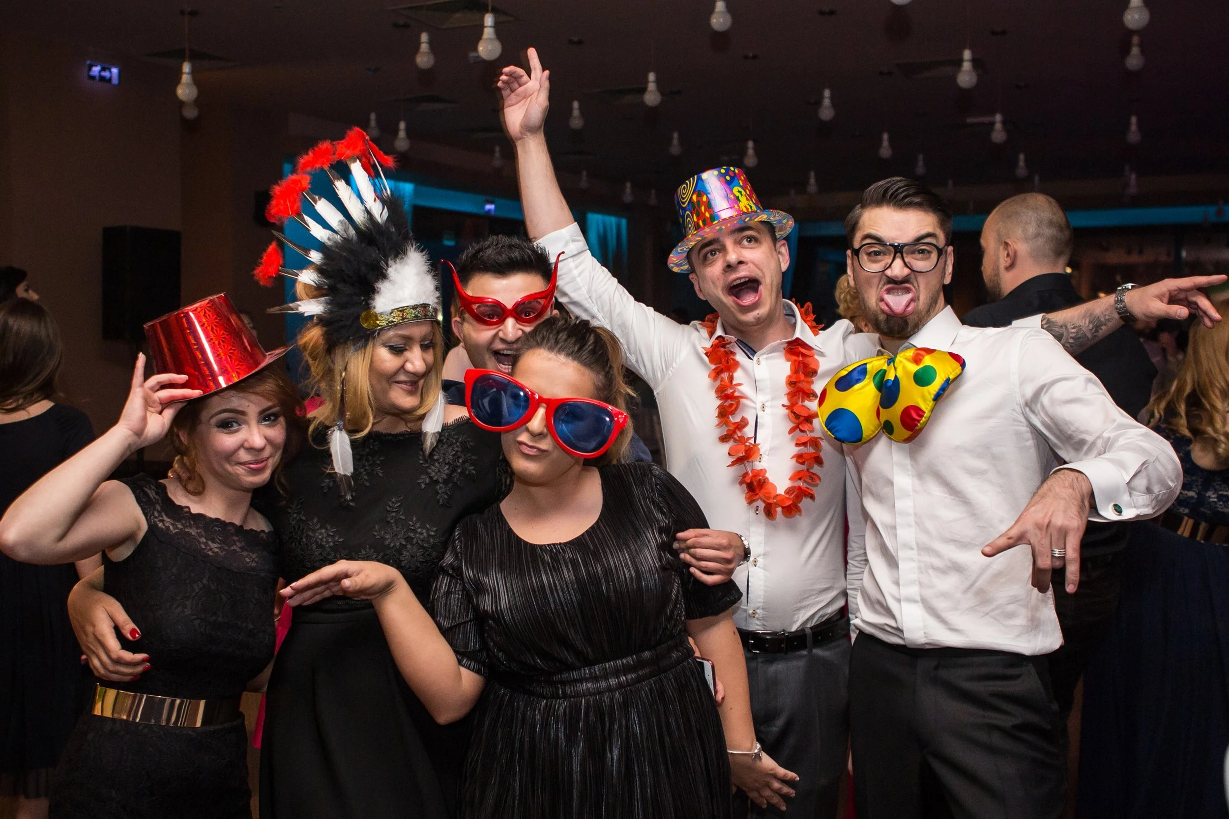 Group of people celebrating at a party, wearing colorful accessories such as a feather headdress, large sunglasses, a striped hat, and a bow tie, with many light bulbs hanging from the ceiling in the background.