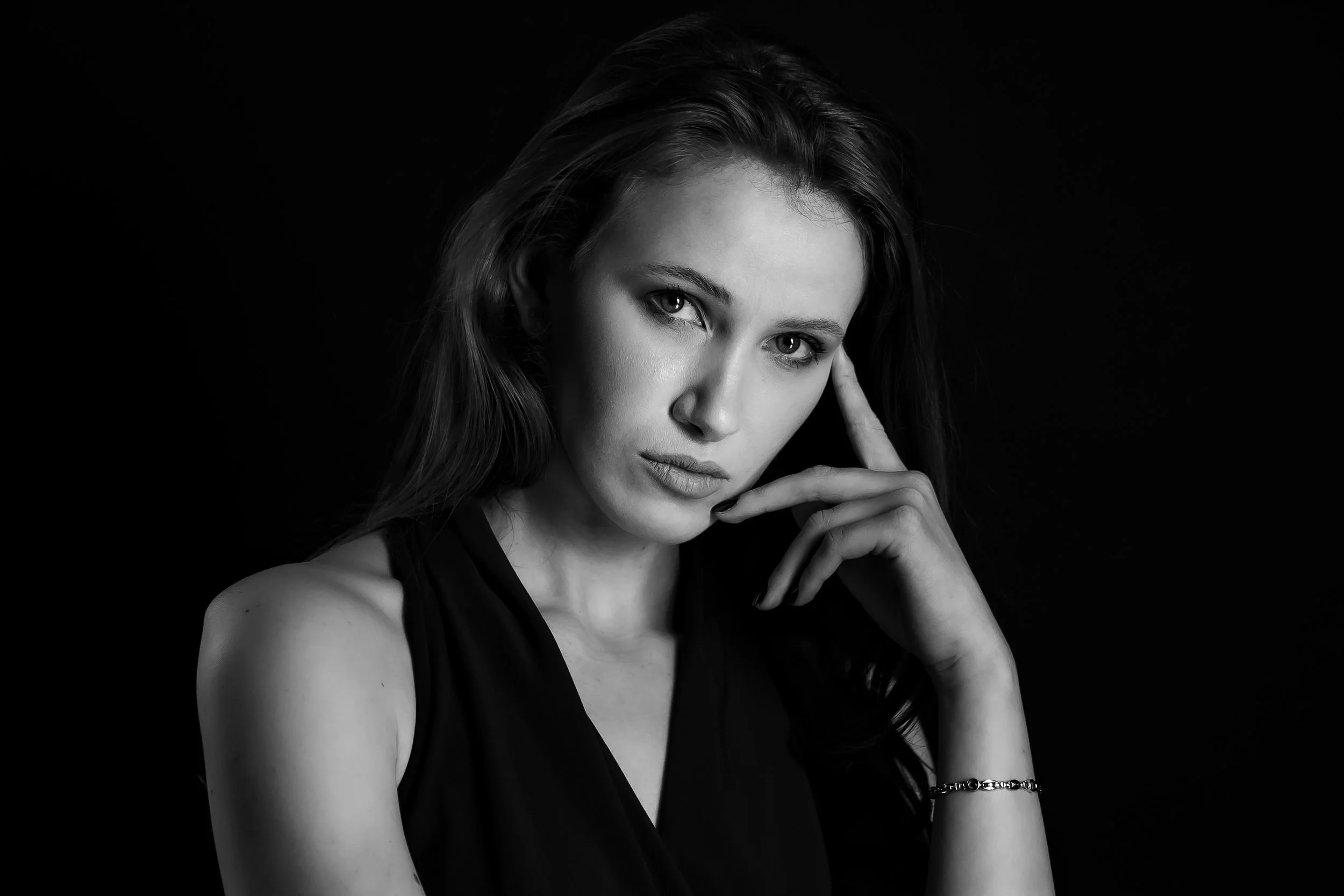 A black-and-white portrait of a young woman with long hair, touching her temple with her right hand, against a dark background.