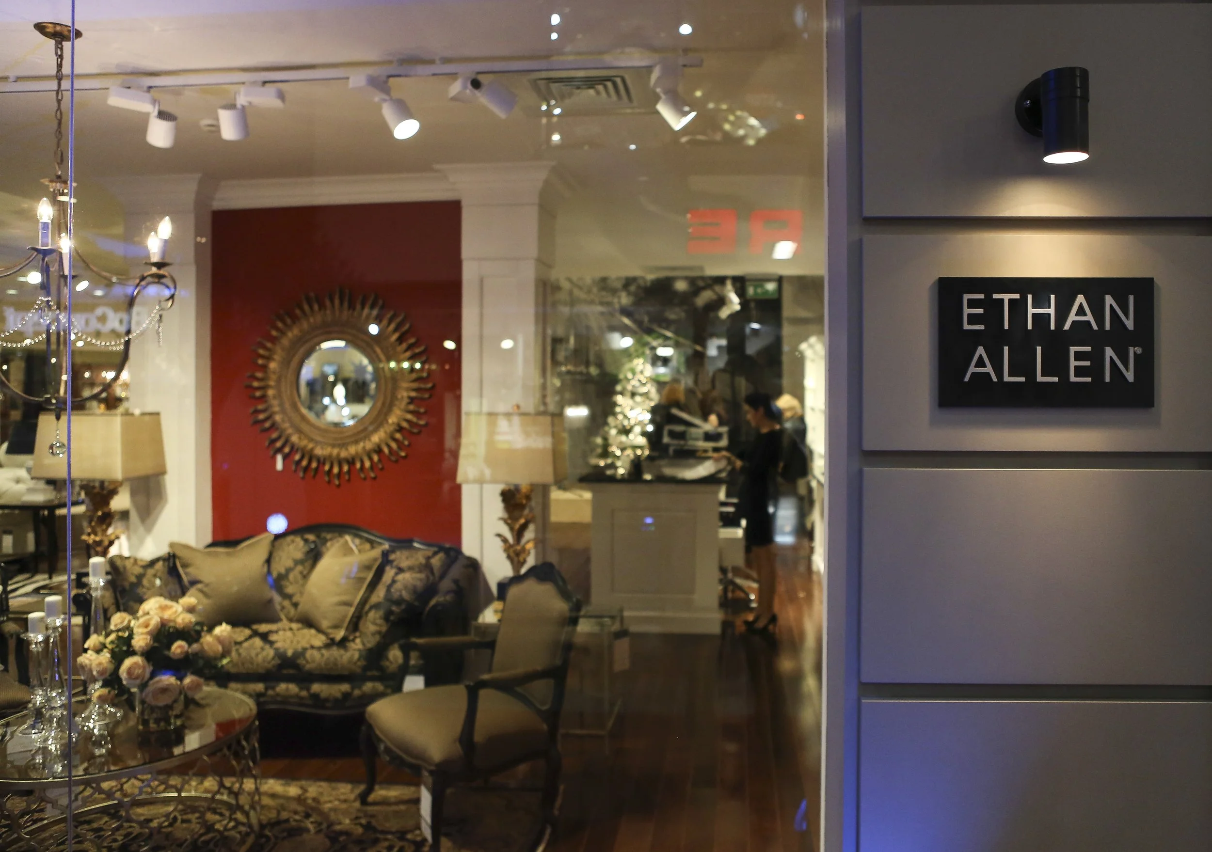 Interior of a furniture store with a sofa, floral arrangement, chandelier, and decorative mirror on red wall, seen through a glass window with a sign for Ethan Allen.