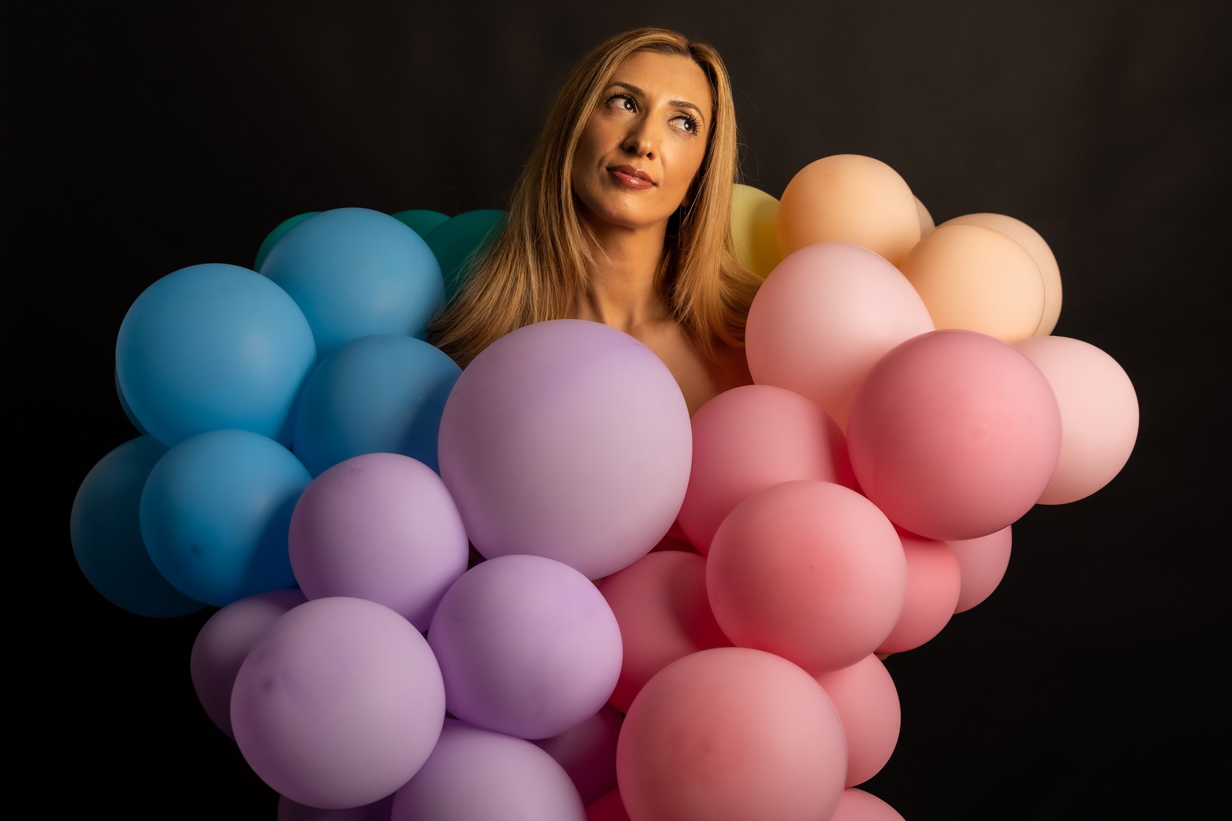 A woman with long blonde hair surrounded by pastel-colored balloons in shades of blue, pink, peach, and purple against a black background.