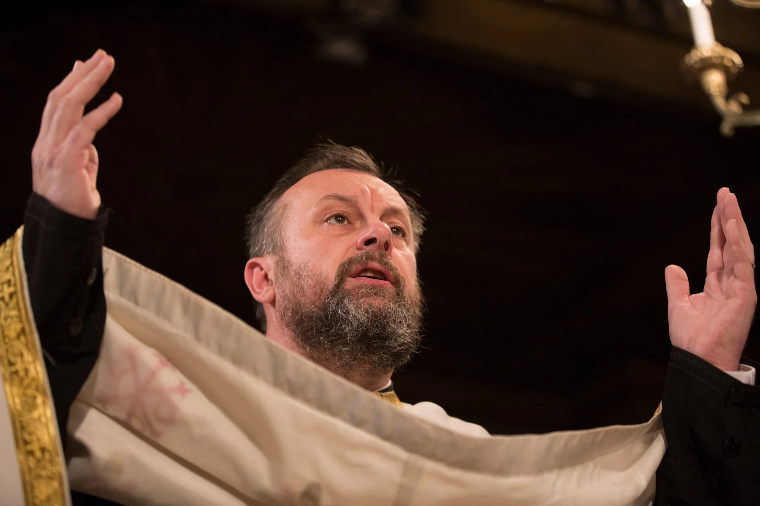 A man with a beard and short hair, wearing a cream-colored gown with gold trim, raises his hands in a gesture of blessing during a religious ceremony, with a dark background and a chandelier overhead.