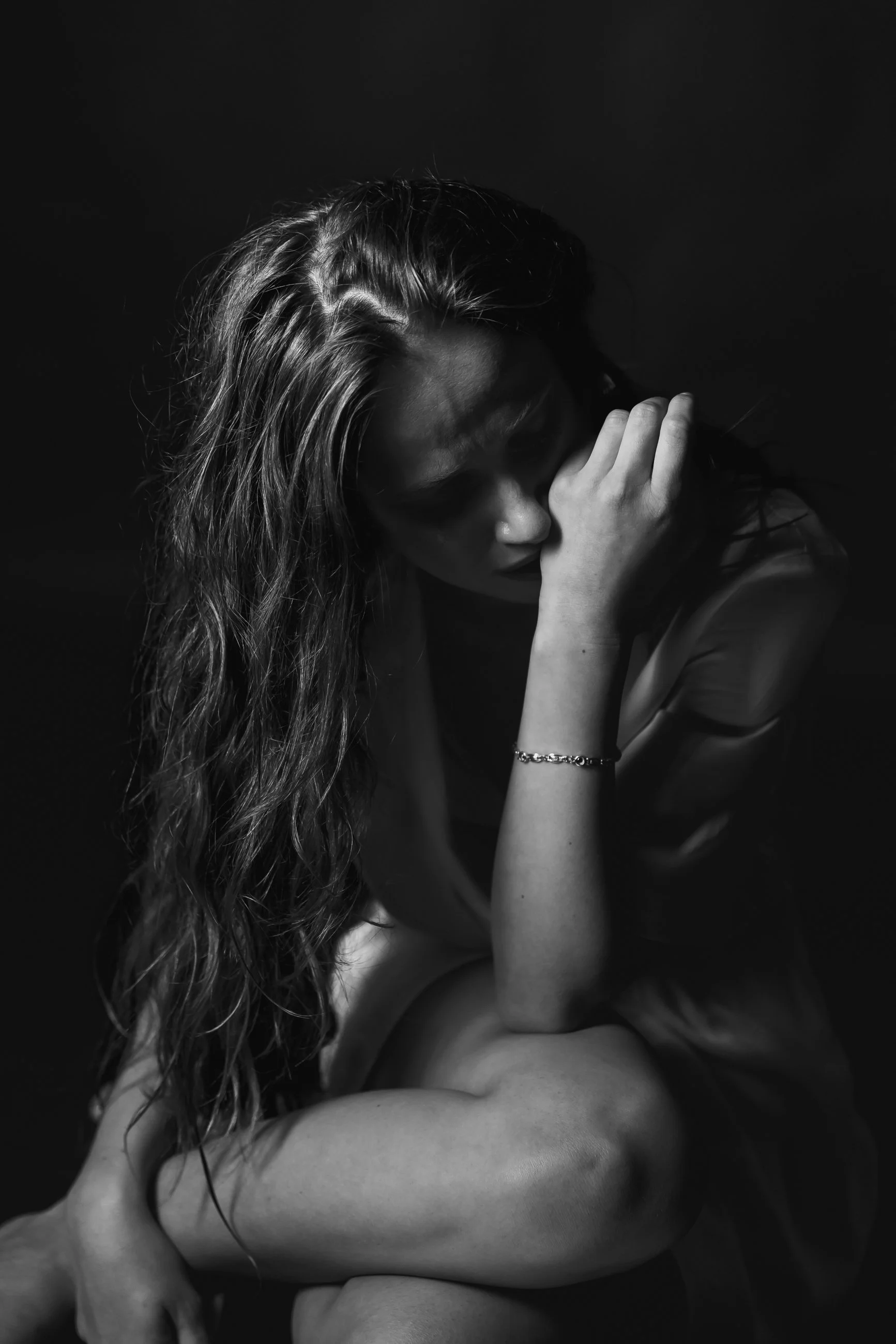 A black and white photograph of a woman with long hair, sitting with her eyes closed, resting her forehead on her hand, and wrapping her arm around her knee.
