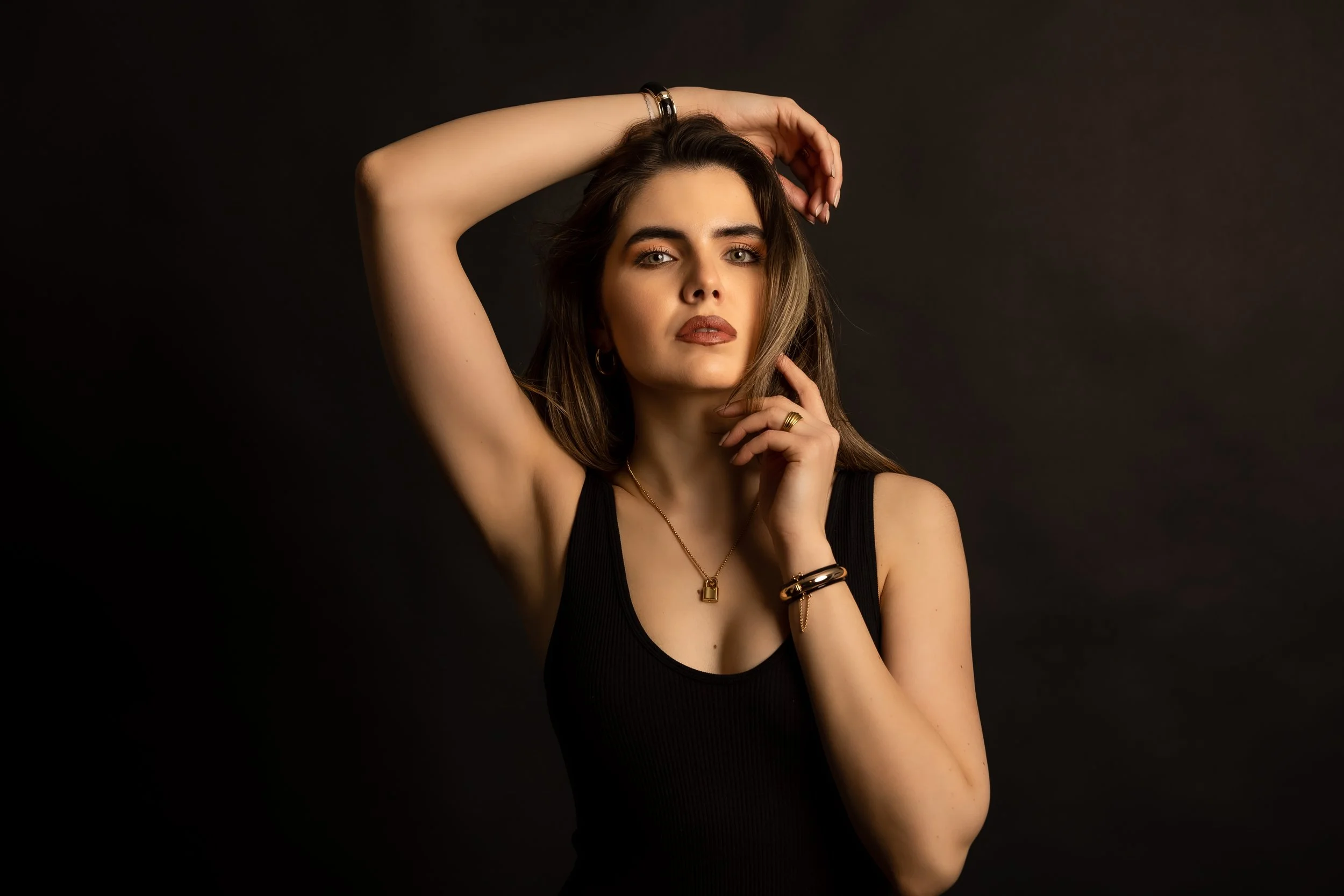 A woman with long brown hair and fair skin poses with her right arm raised and her head resting on her hand against a dark background. She is wearing a black sleeveless top, jewelry including earrings, rings, bracelets, and a necklace, and makeup wit