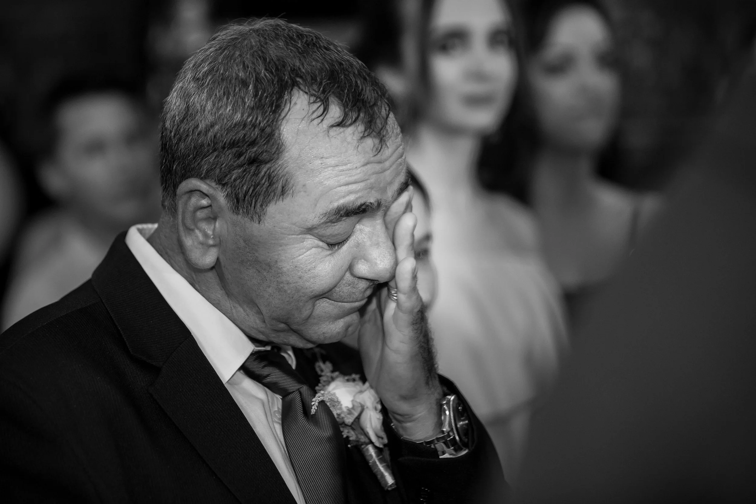 A man in a suit and tie with a wedding corsage is holding his hand to his face in an emotional moment during a formal event, with blurred women in the background.
