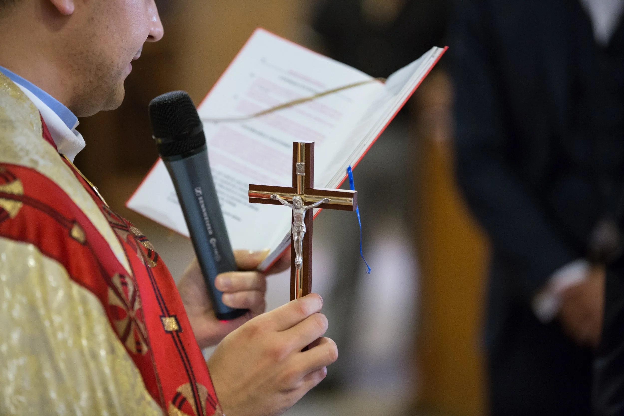 A person holding a microphone and a book with a crucifix in front of them, likely during a religious ceremony or service.