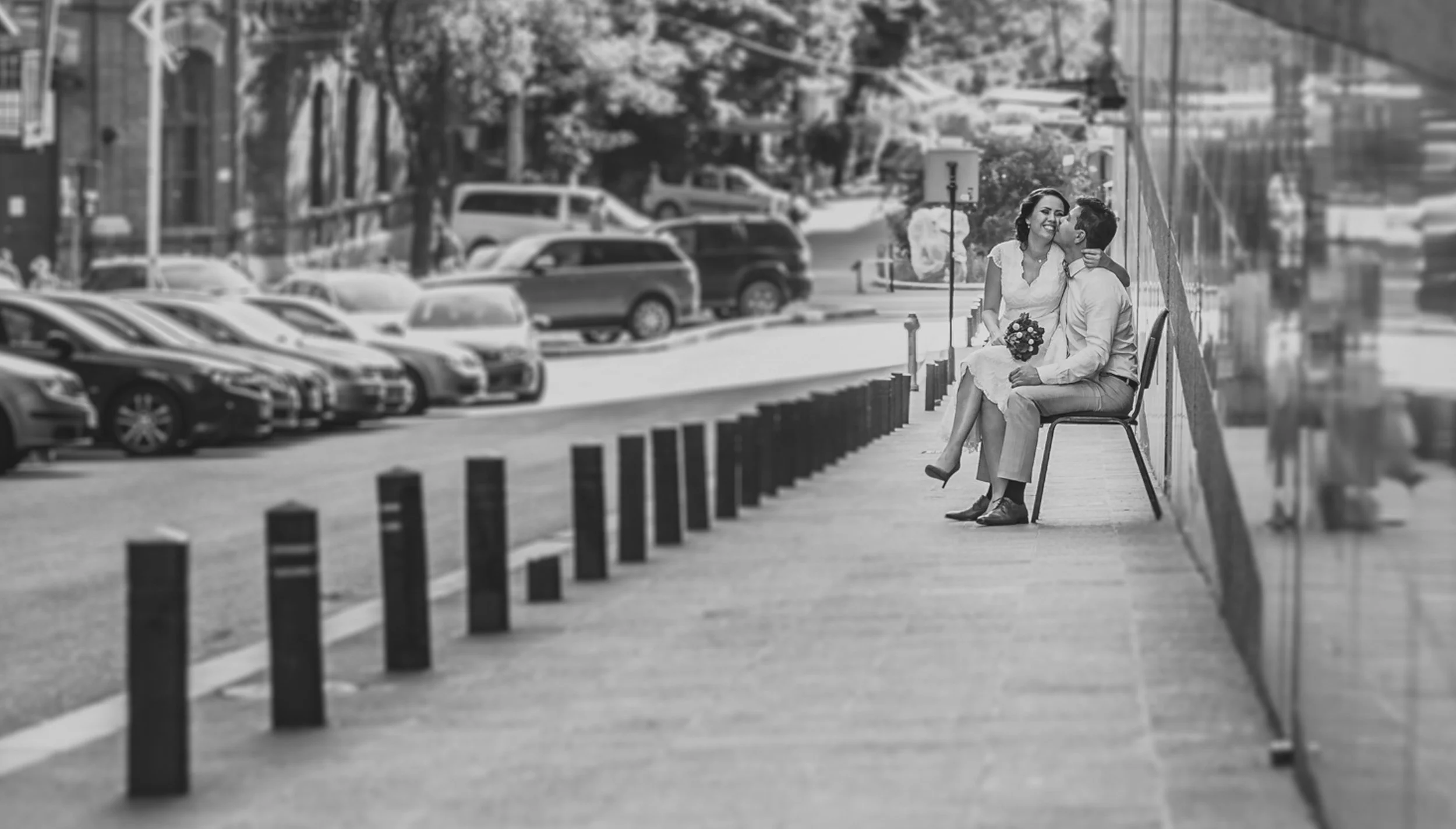 A couple sitting on a bench on a city sidewalk, the woman holds a small bouquet of flowers and kisses the man's cheek while he looks up at her, with parked cars and trees in the background.
