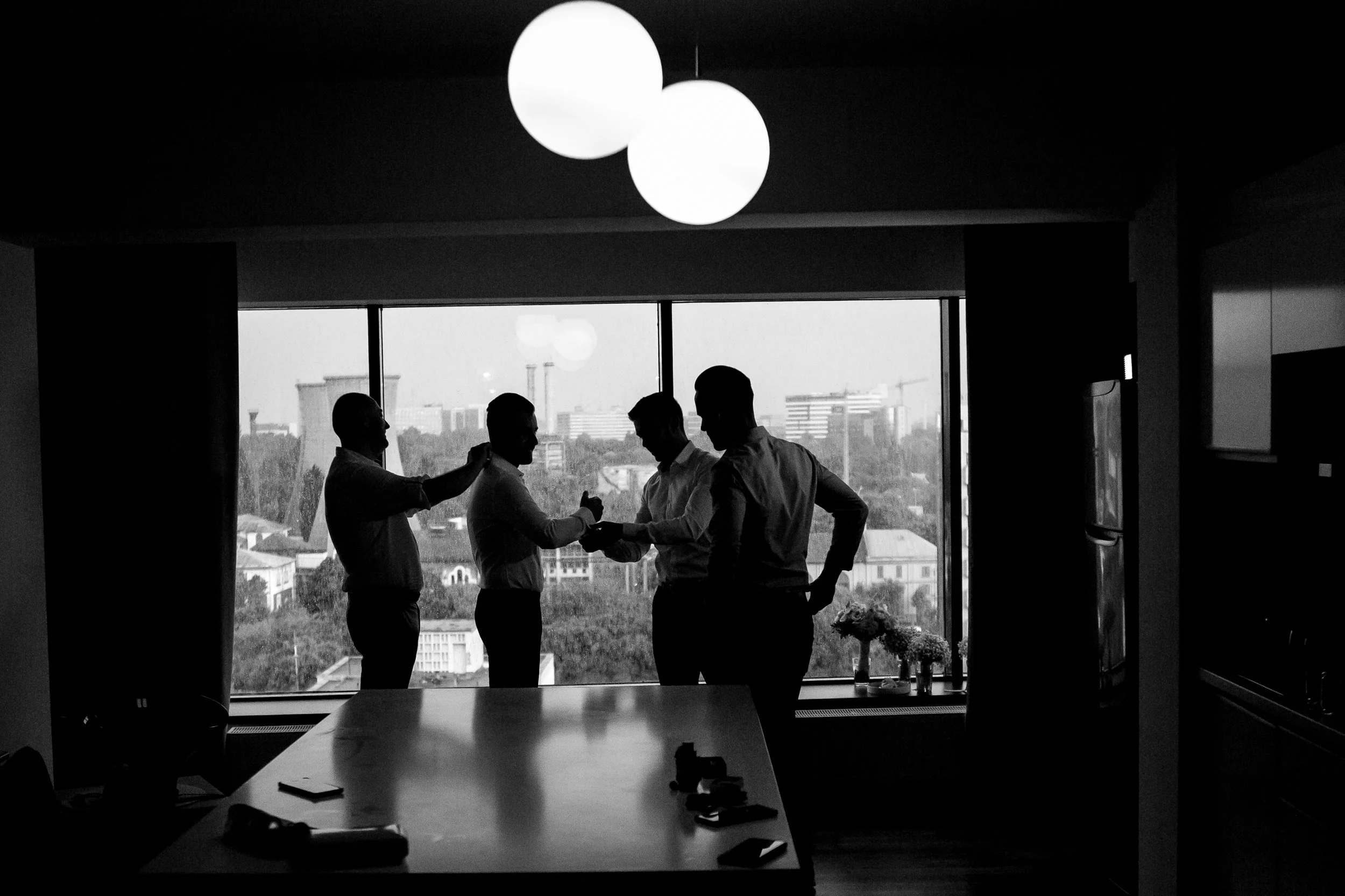 Four men in business attire having a discussion in front of a large window overlooking a cityscape, with a bright light fixture hanging from the ceiling in a modern office setting.
