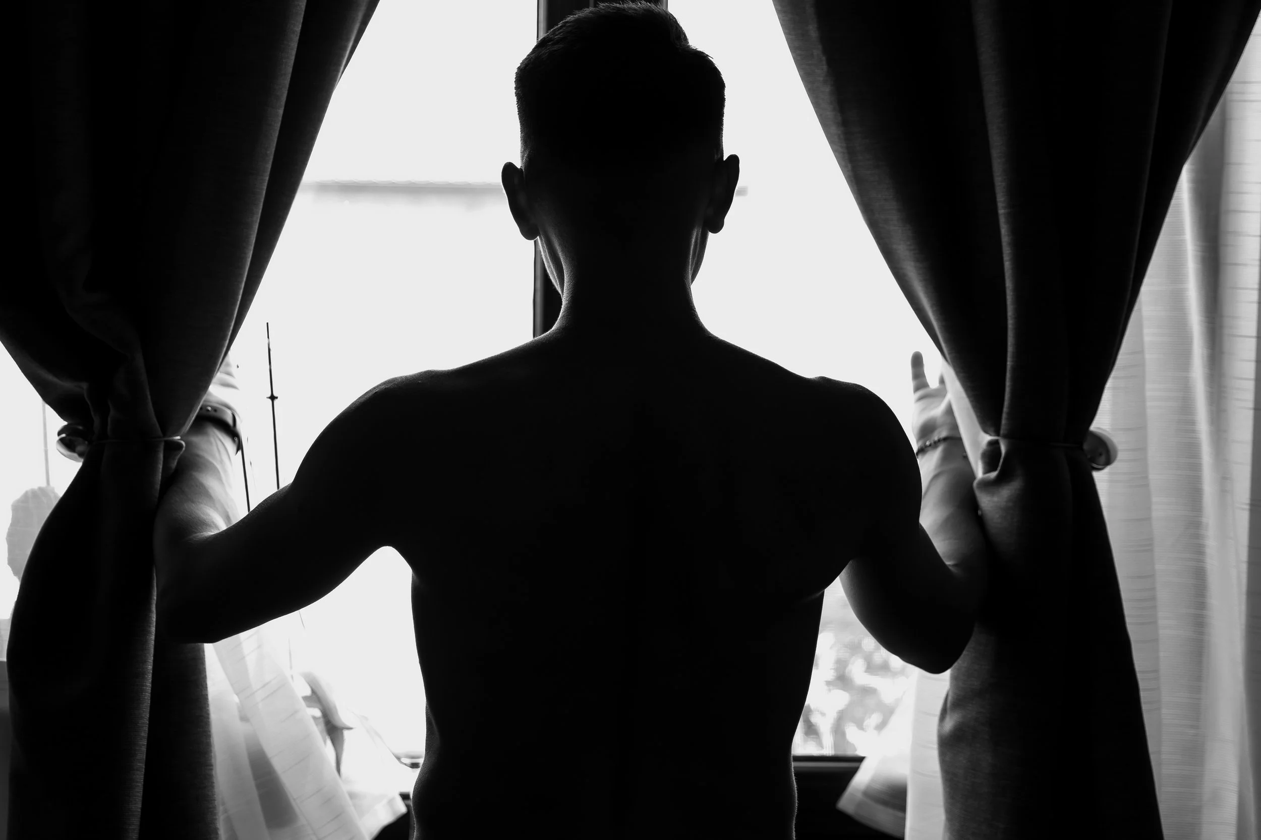 Silhouette of a person standing in front of a window with curtains, adjusting the curtains, in black and white.