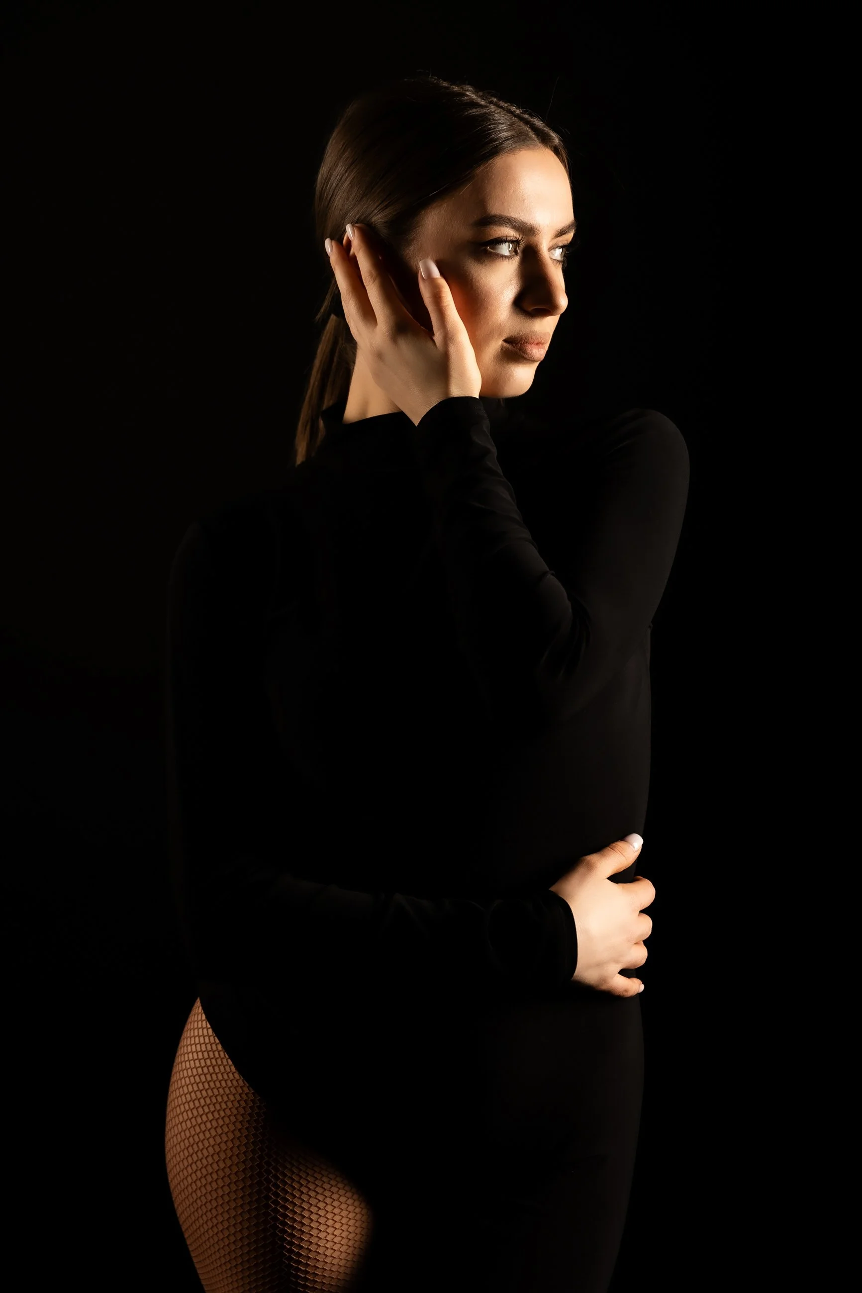 A woman with long brown hair in a ponytail, wearing black long-sleeve top, fishnet stockings, and posing against a black background with her hand on her face and her other arm crossed over her waist.