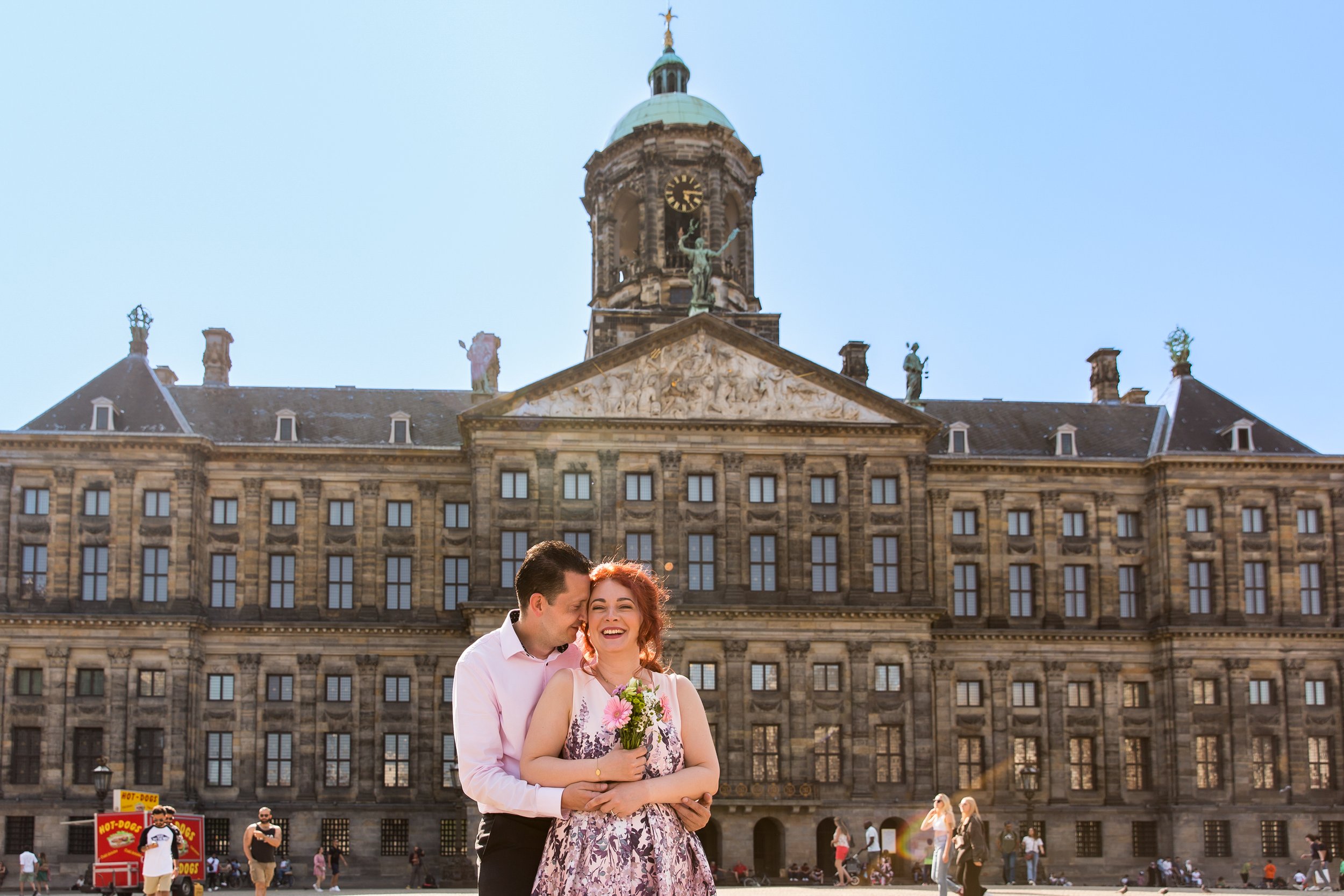A couple stands close together in front of a historic building with a clock tower, holding a bouquet of pink flowers, smiling and enjoying a sunny day. People walk in the background, and there is a hot dog stand nearby.