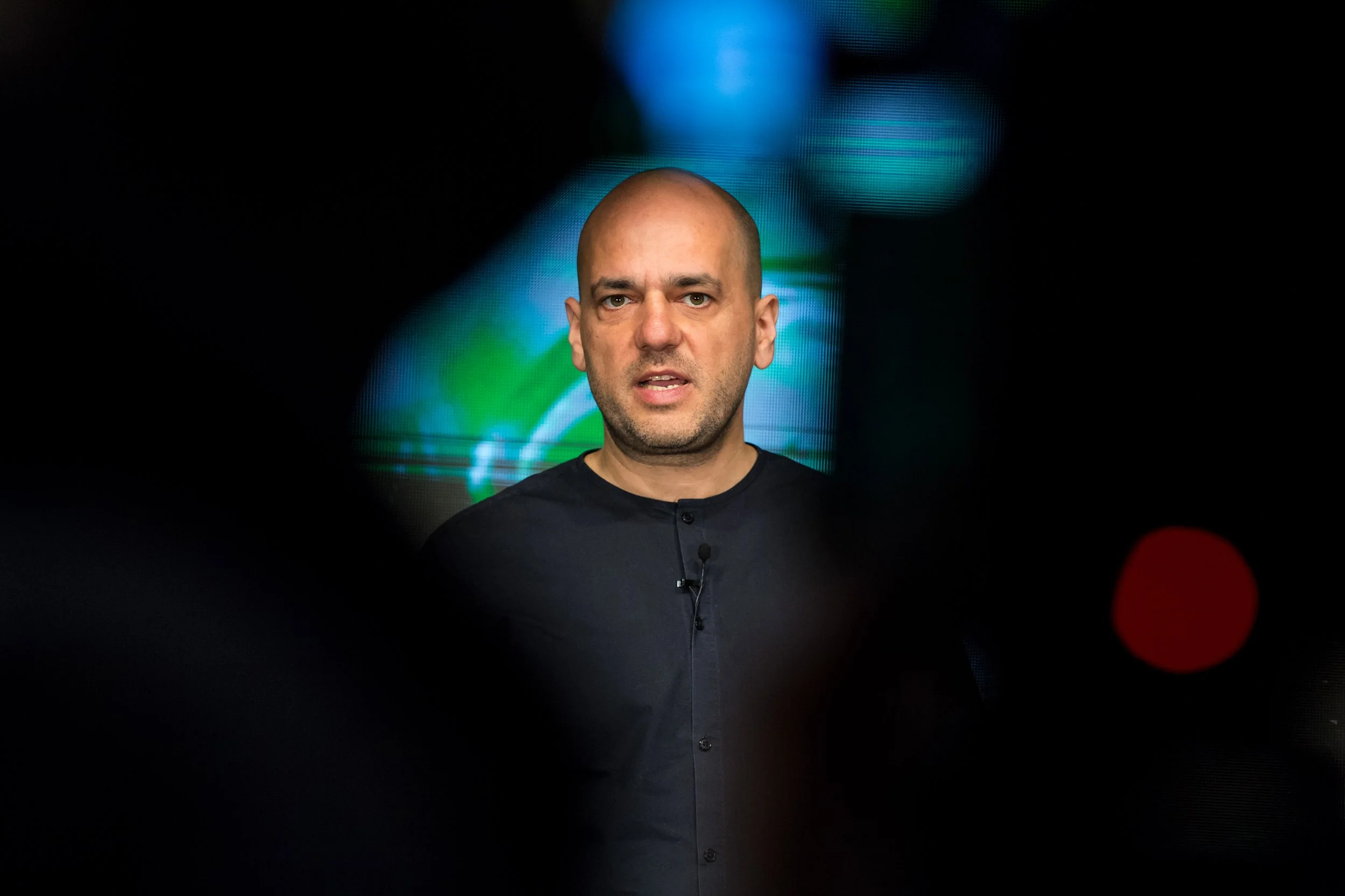 A man with a bald head and light skin, wearing a black shirt, speaking during an event. The background shows a blurred screen with colorful graphics, and the image is framed by out-of-focus dark and red elements.