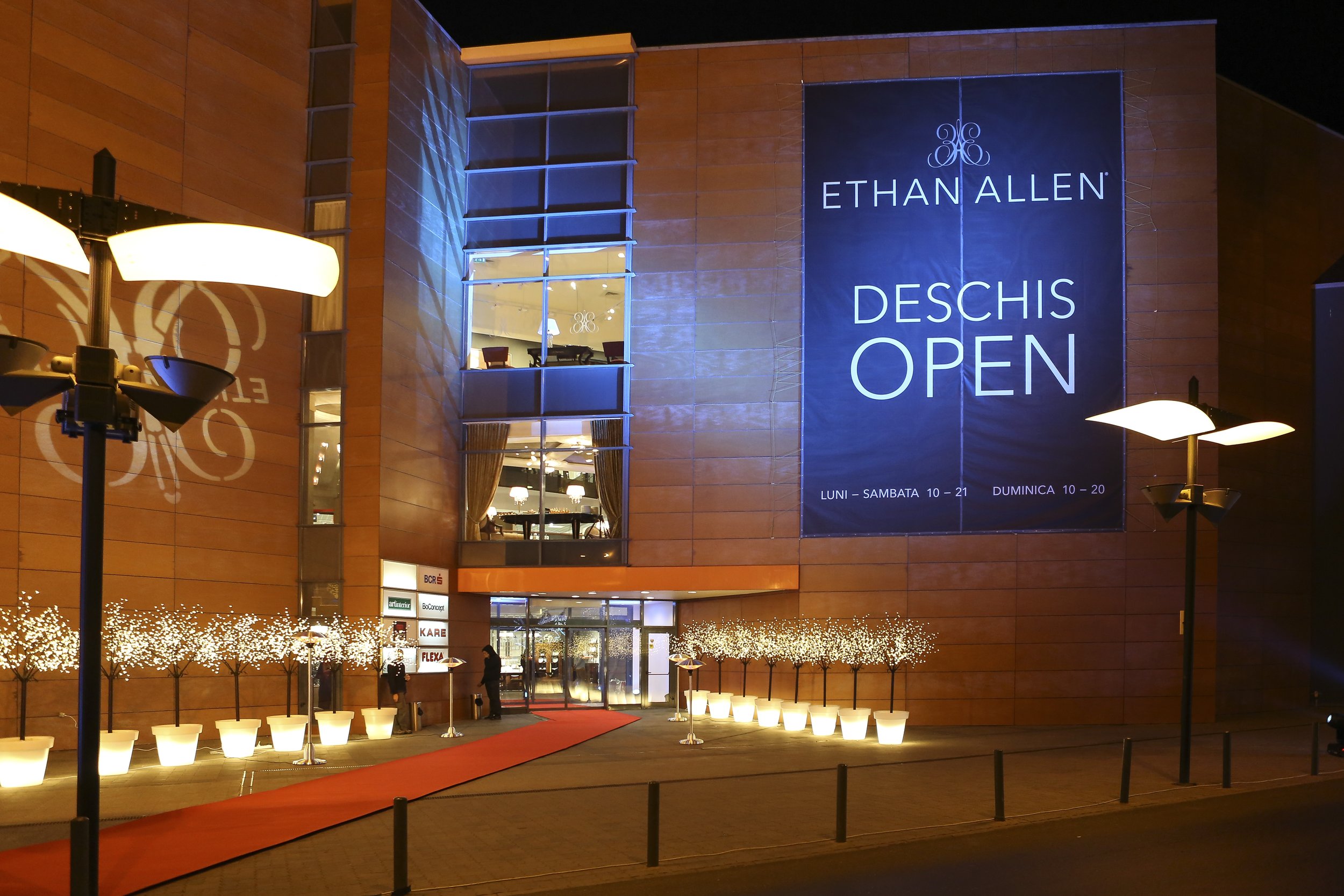 Night view of a modern building with glass windows and a large promotional banner that reads 'Ethan Allen Deschis Open' with business hours listed below. The entrance is decorated with white illuminated planters, a red carpet, and surrounding trees w