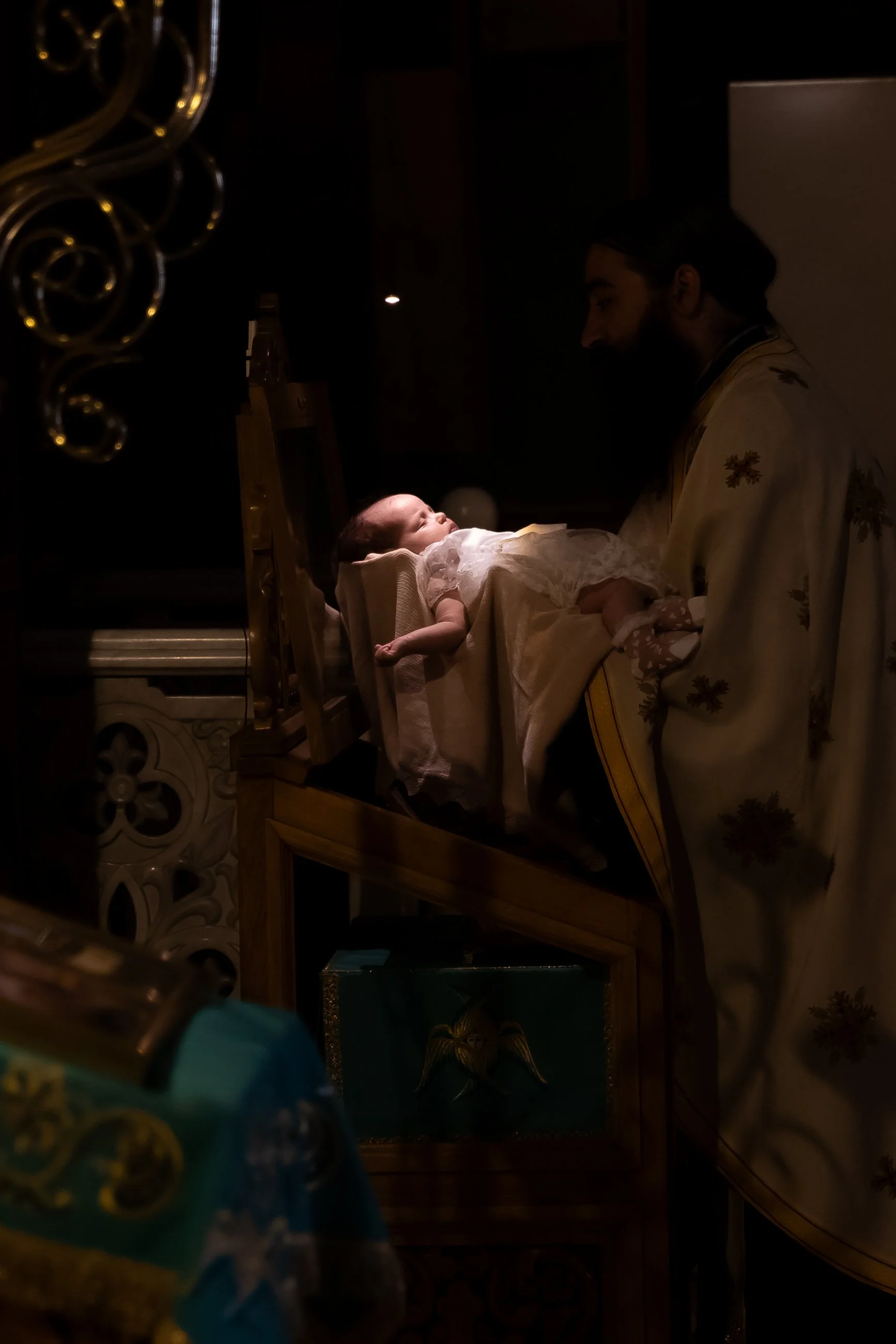 A baby cradled in a man’s arms during a baptism ceremony in a church, with candle holders and religious items nearby.