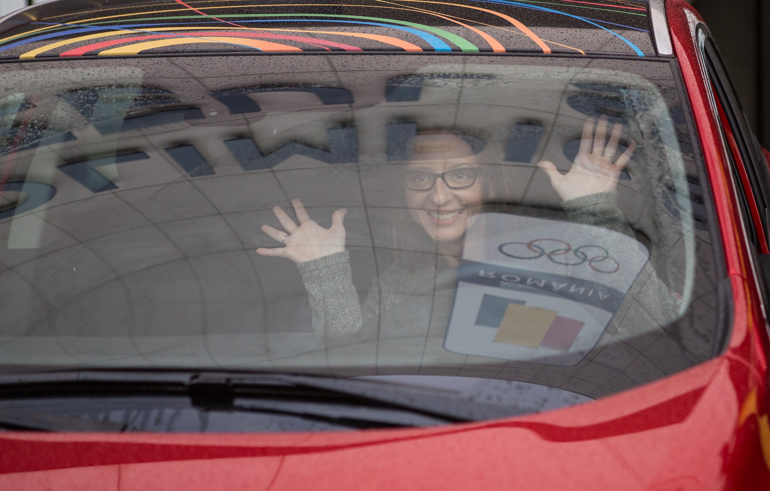 A person with glasses waving and smiling inside a red car, seen through the front windshield with water droplets, a sign with the text 'GESP' and some colorful rainbow designs on the roof.