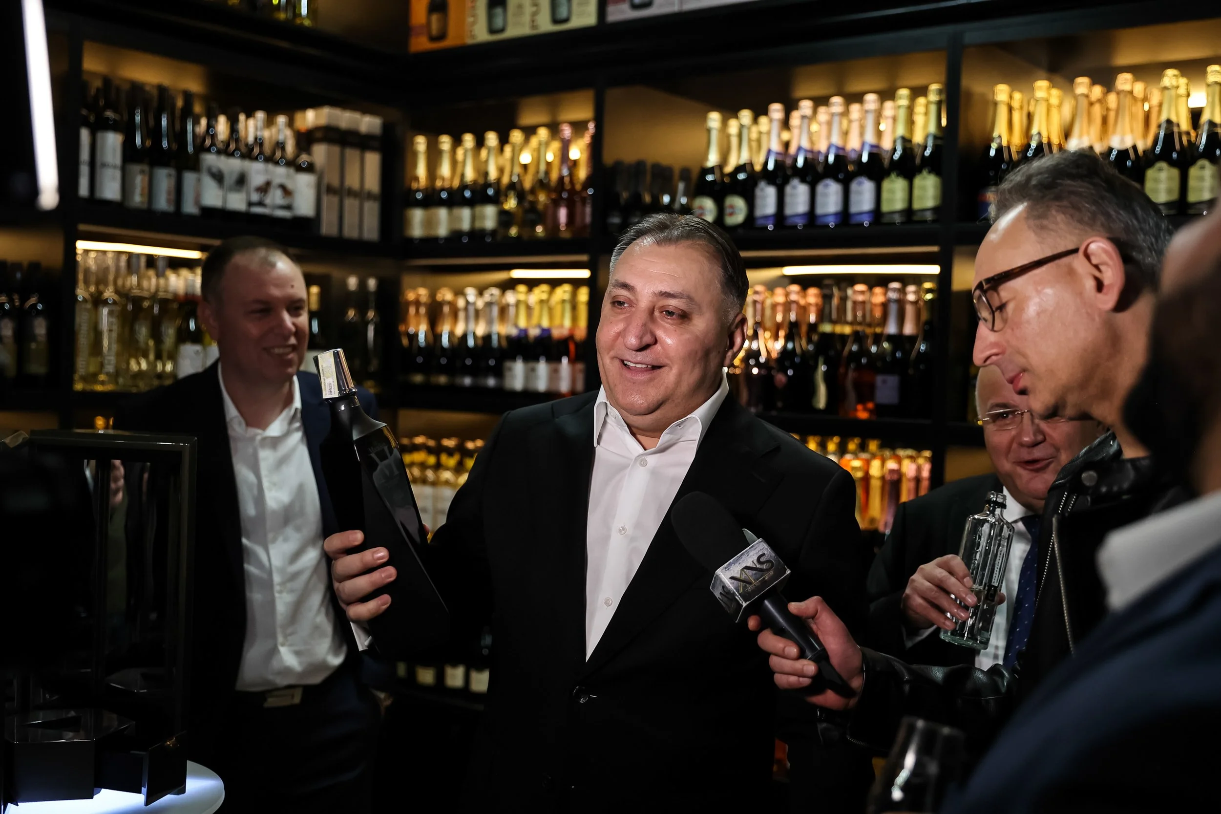 People in suits attending a wine tasting event with shelves of wine bottles in the background, one person holding a wine bottle and microphones present.