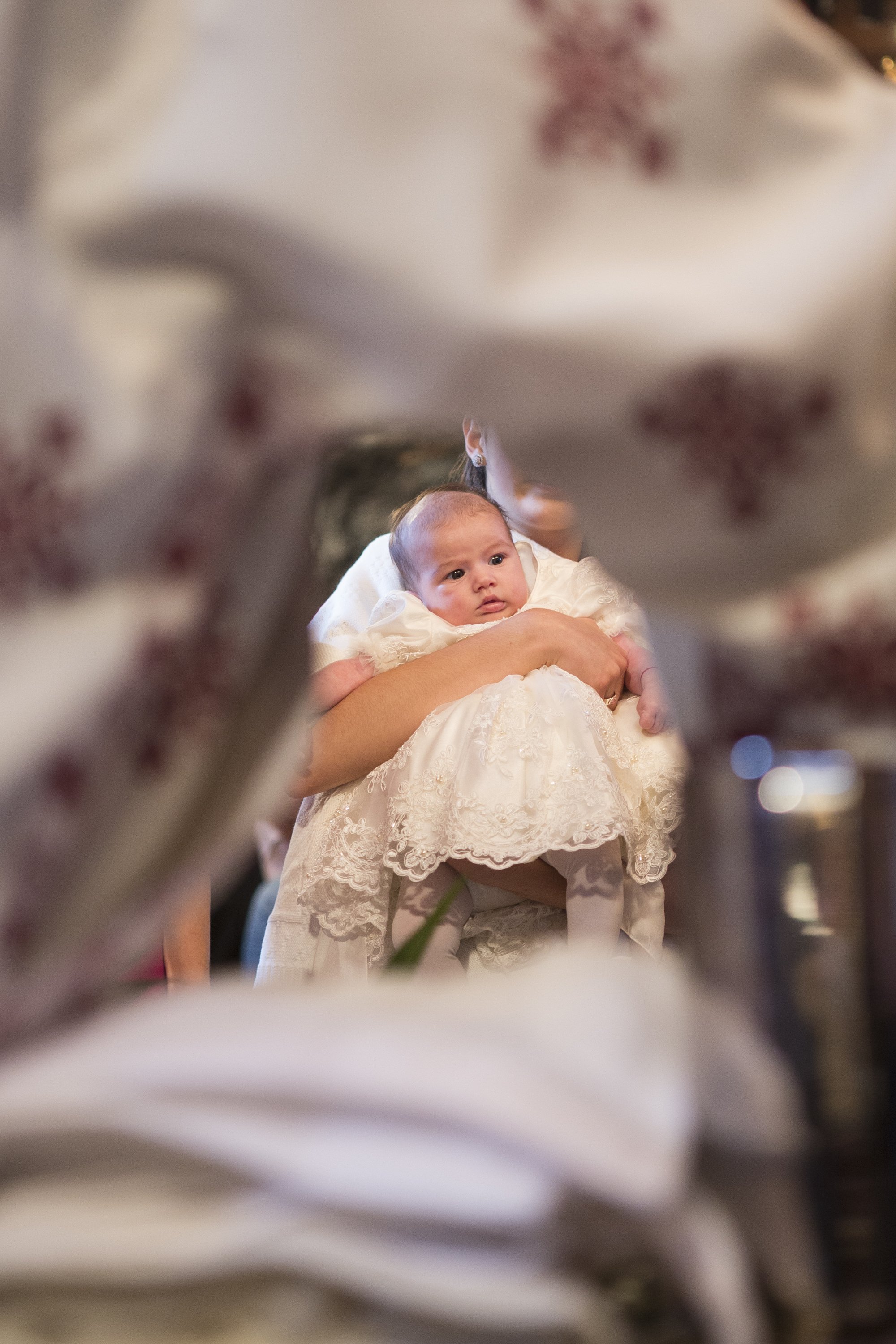 A baby being held up during a baptism or christening ceremony, dressed in a white gown, viewed through a floral or decorative frame.
