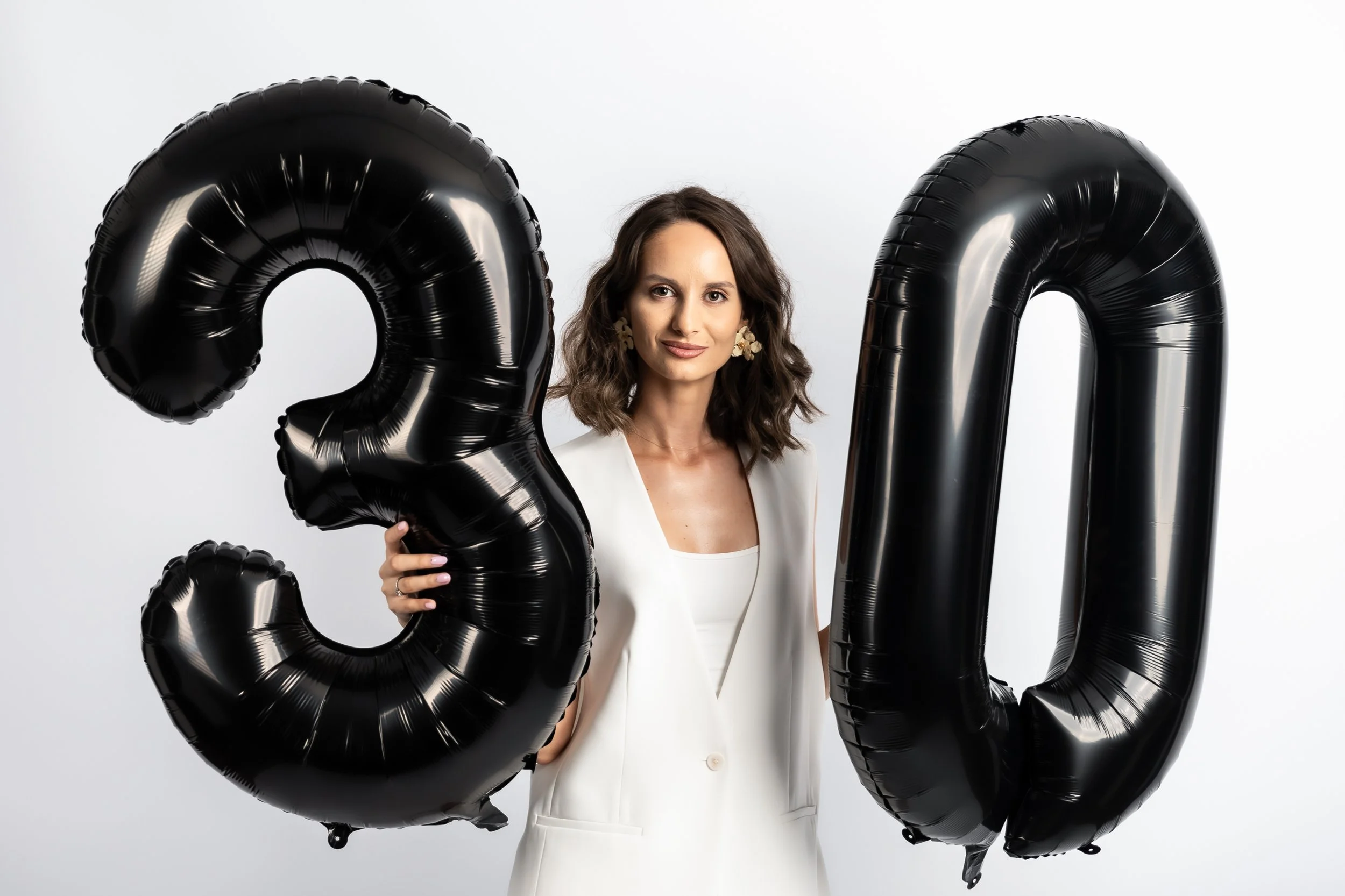 A woman with wavy brown hair wearing a white blazer and earrings holding large black balloons shaped like the number zero and the number two.