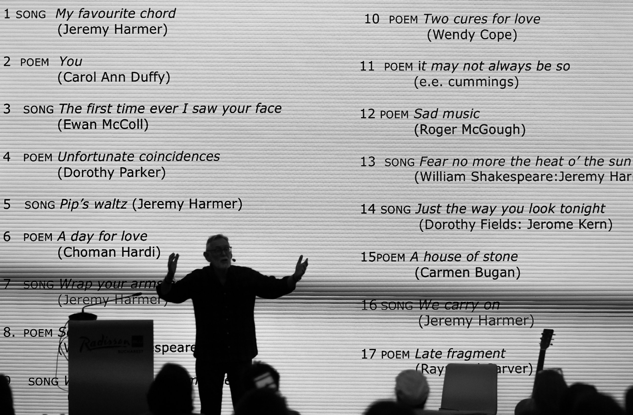 A person standing in front of a large sheet of paper with a numbered list of titles and authors of poems and songs, possibly during a presentation or reading, with an audience in the foreground.