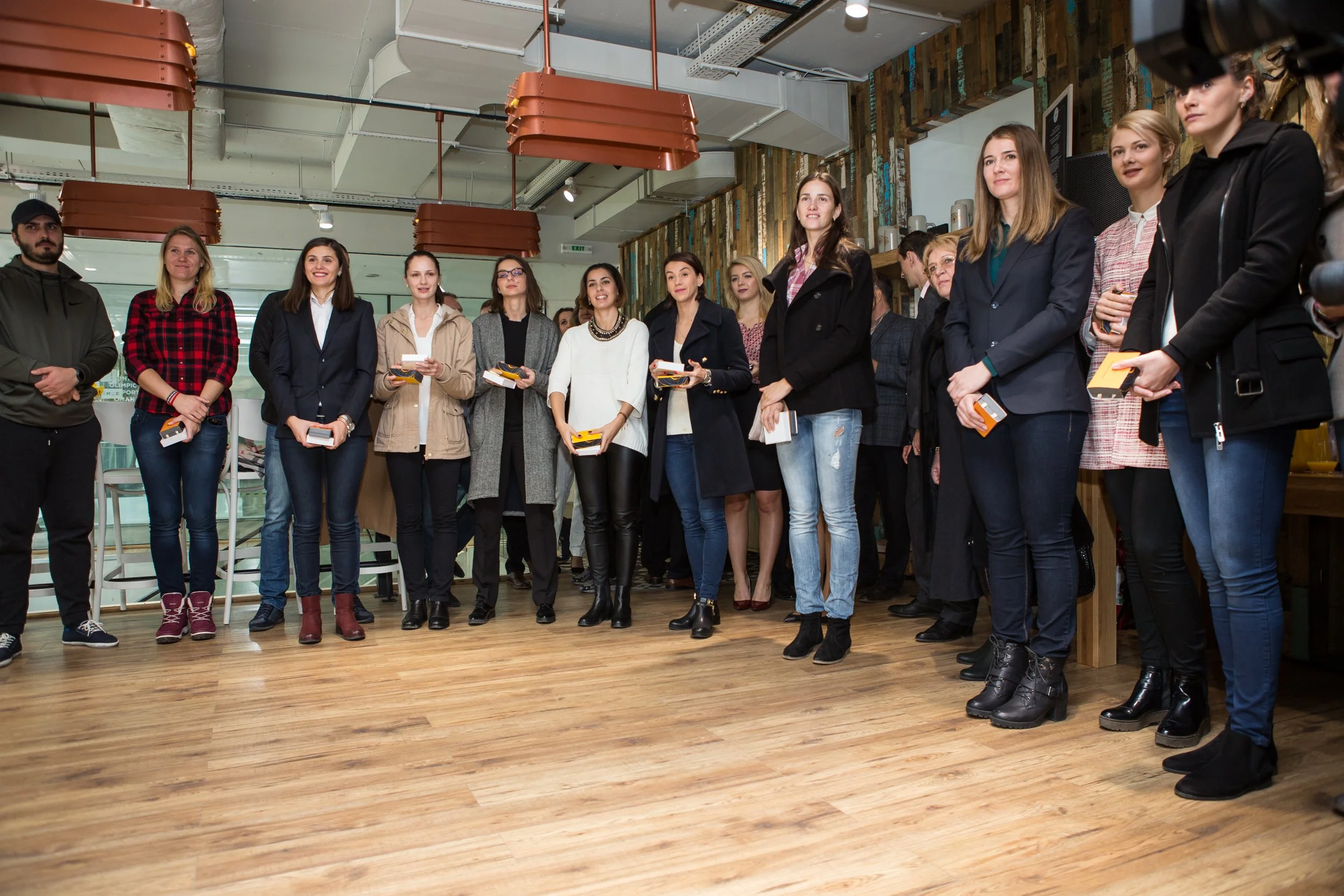A group of diverse women standing in a line, some holding small boxes or notebooks, in a modern indoor space with wooden flooring and contemporary decor.