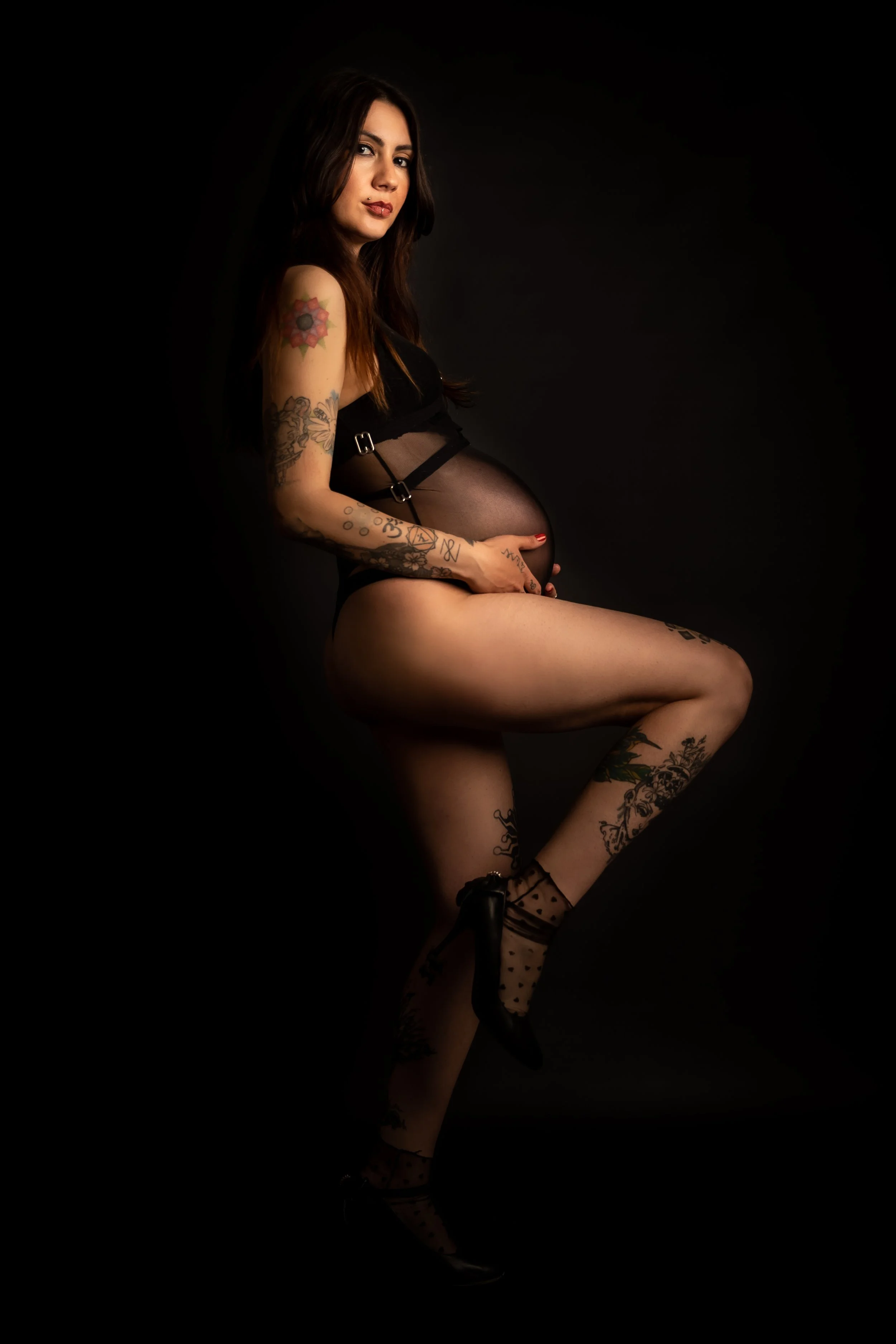 A woman with long dark hair and tattoos on her arms and legs, wearing black lingerie and high heels, sitting against a black background.