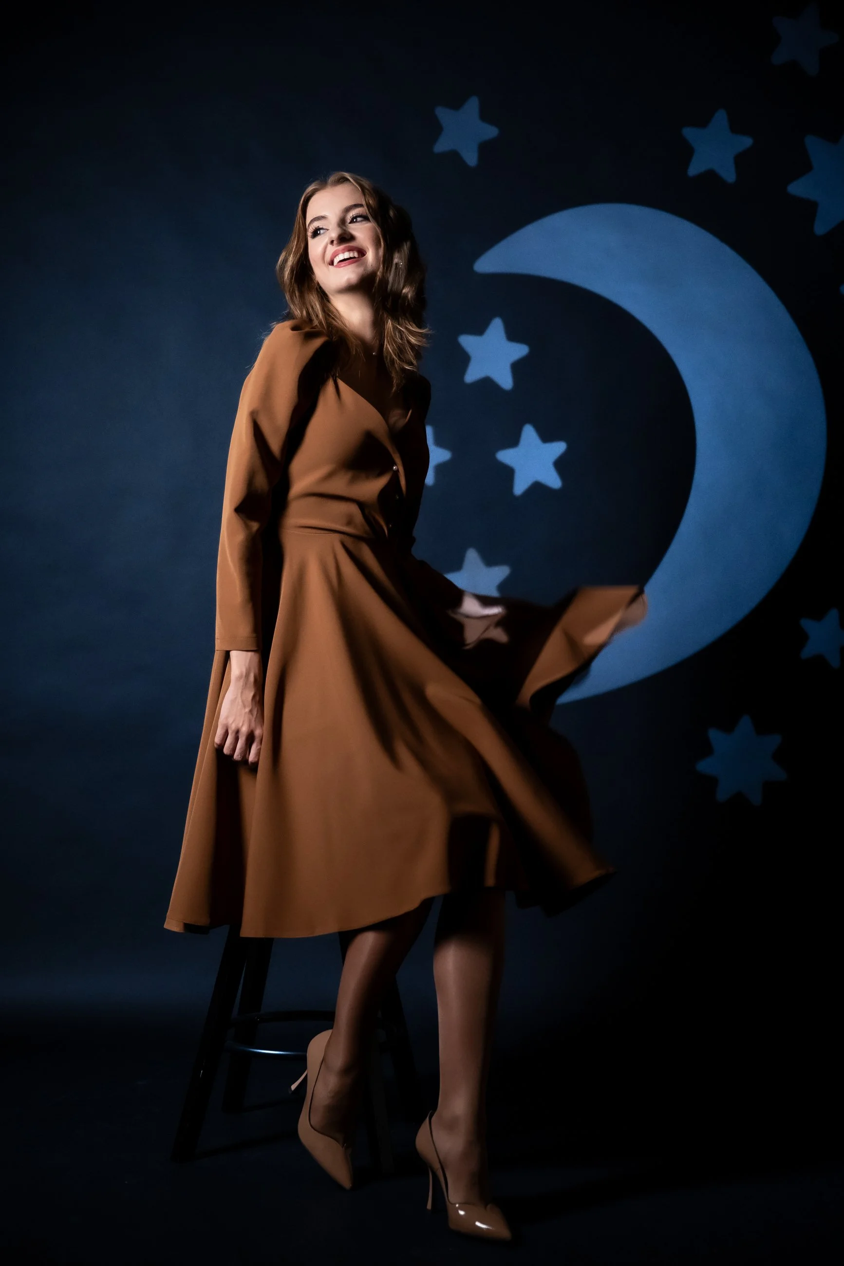 A woman with wavy brown hair in a brown dress and high heels sitting on a stool with a dark background featuring a large blue crescent moon and blue stars.