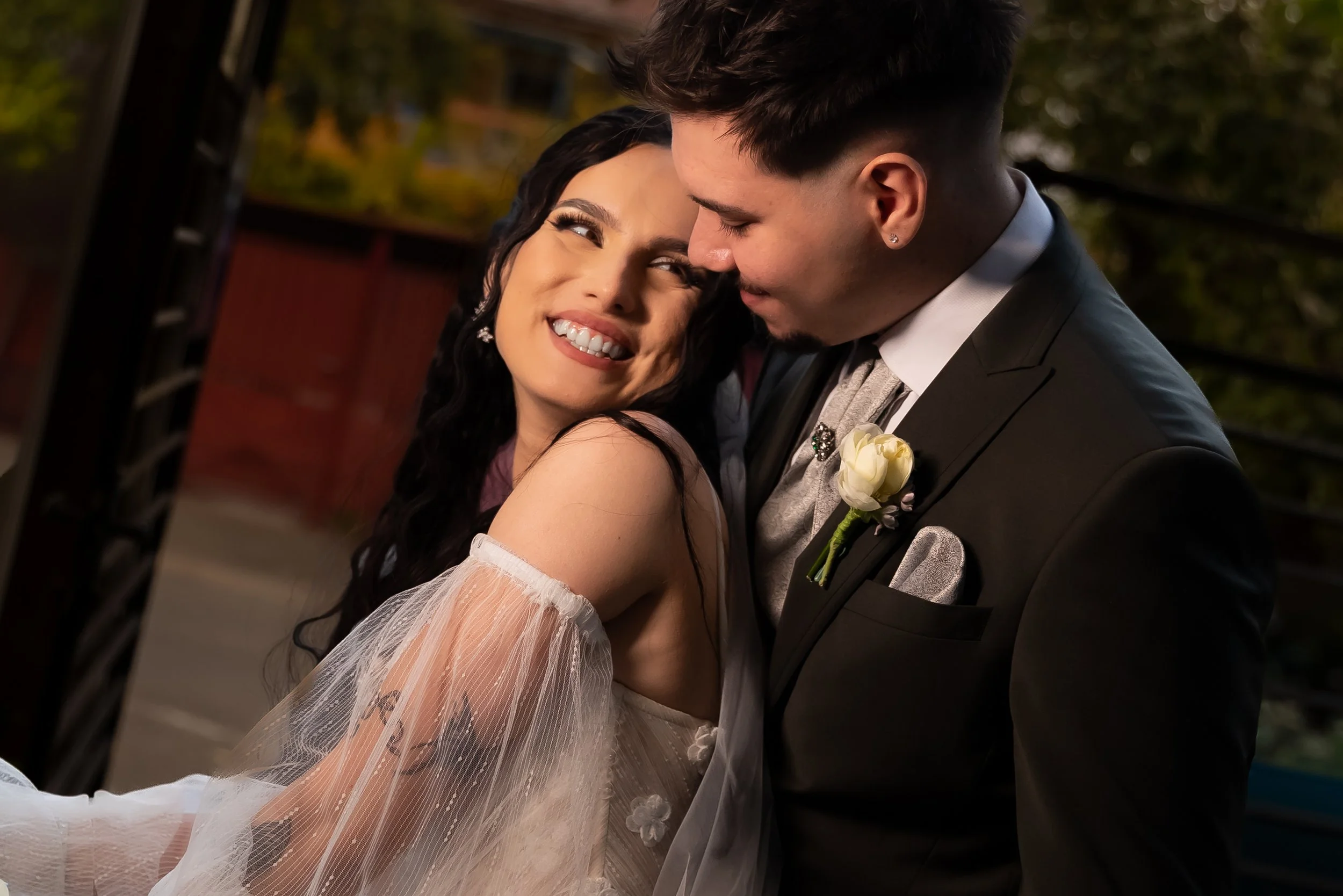 A smiling bride and groom sharing a close moment at their wedding, outdoors with a blurred background of trees.