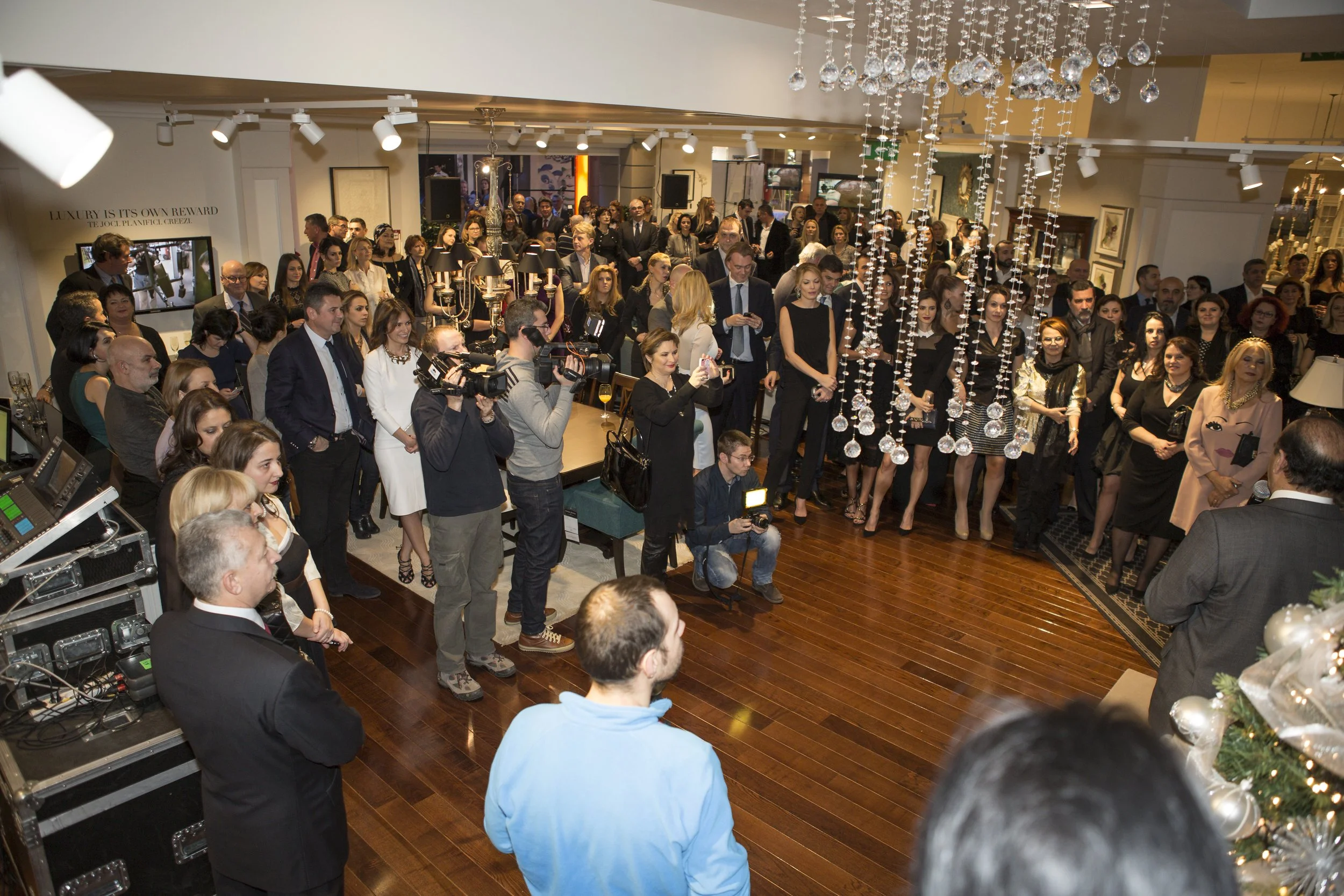A large gathering of well-dressed people attending an event or party in a stylish interior with hanging crystal decorations, lighting, and framed artwork on the walls.
