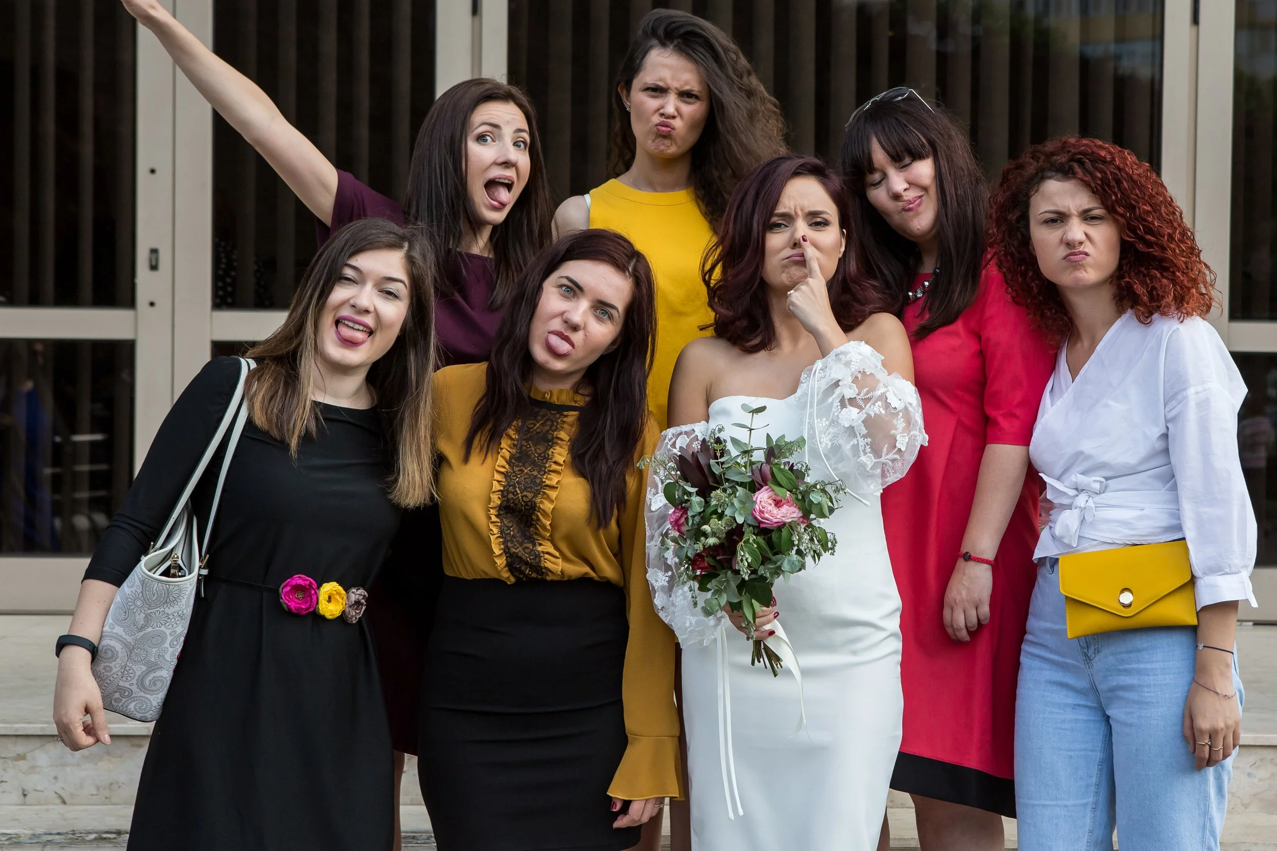 A group of seven women posing together outdoors, one wearing a wedding dress holding a bouquet, and others making playful faces.