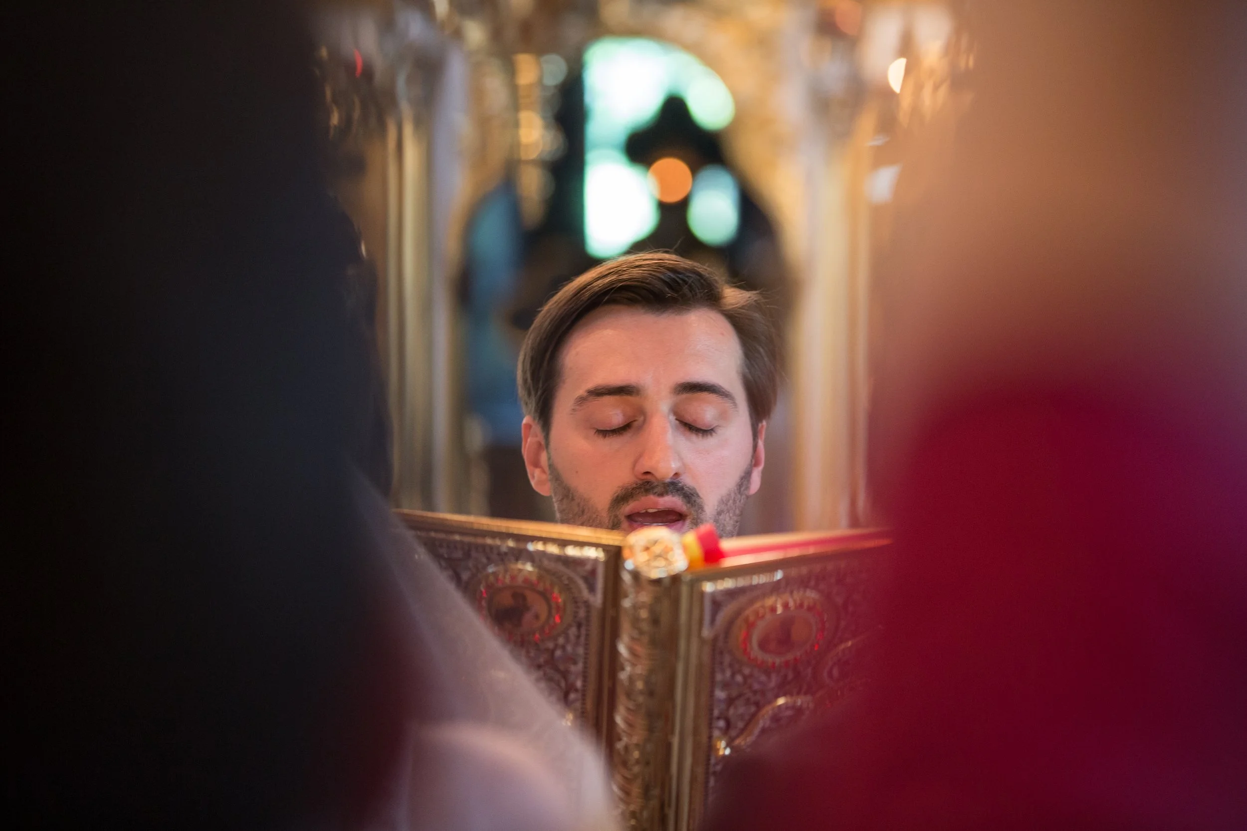 A woman with short brown hair and closed eyes is looking into a mirror. Her reflection is visible in the mirror, which also shows part of her face. The background is blurred with warm lighting, and there is a decorative object with ornate patterns in
