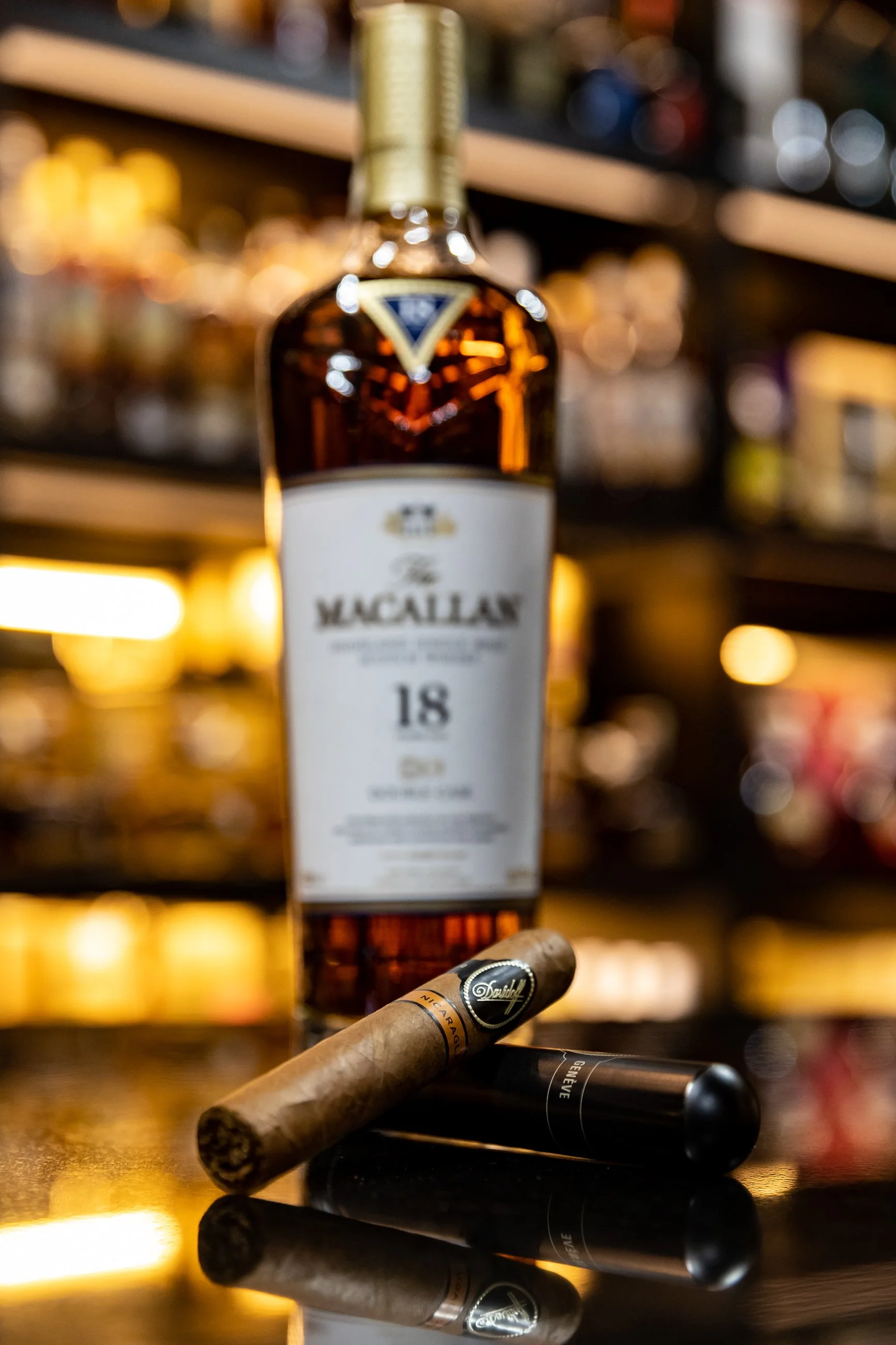 Blurred image of a bottle of Macallan 18 Year Old single malt Scotch whisky on a shelf, with cigars and a cigar cutter in the foreground, against a background of bar bottles and lights.