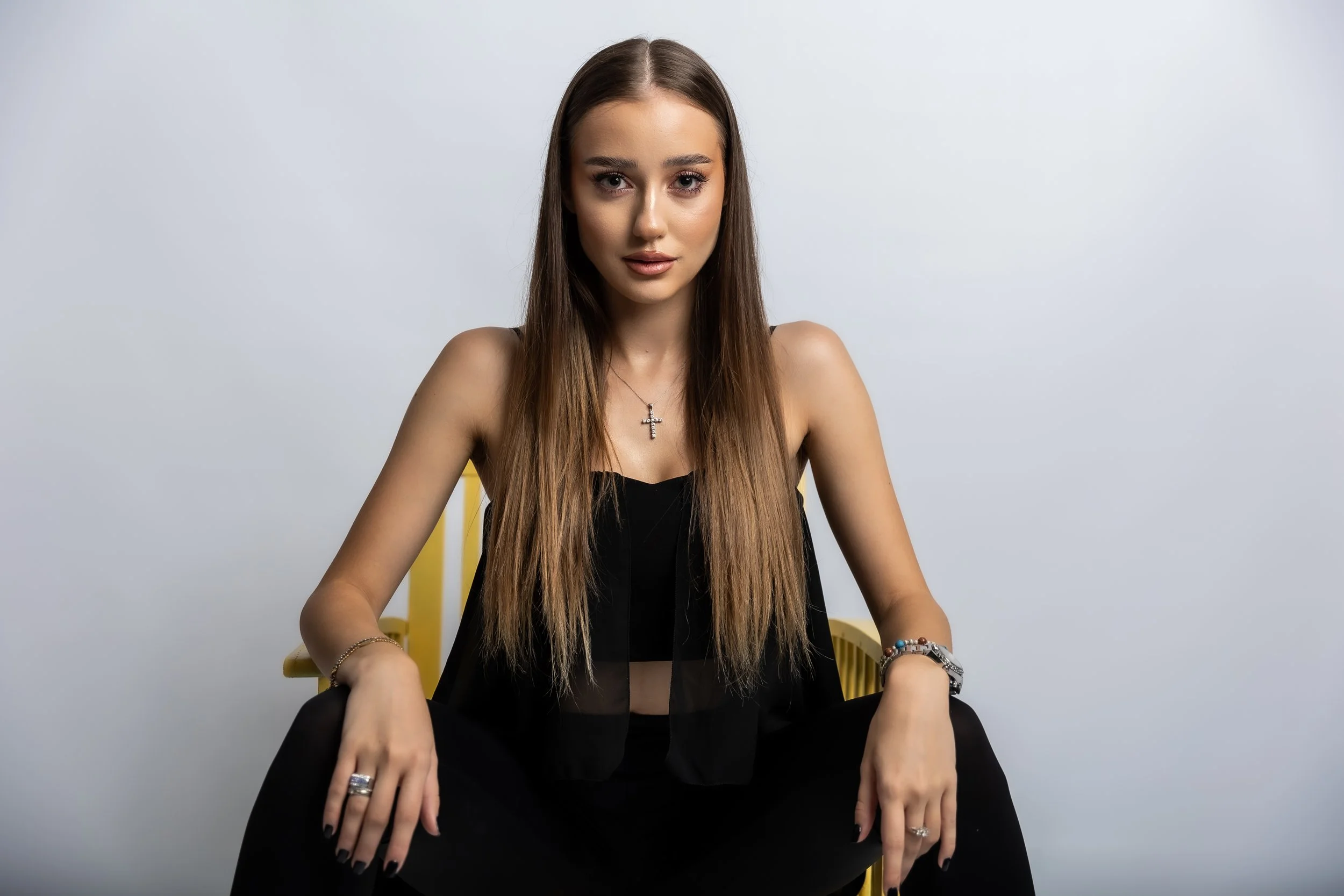 A young woman with long brown hair, wearing a black top, silver cross necklace, and bracelets, sitting on a yellow chair against a plain light gray background.
