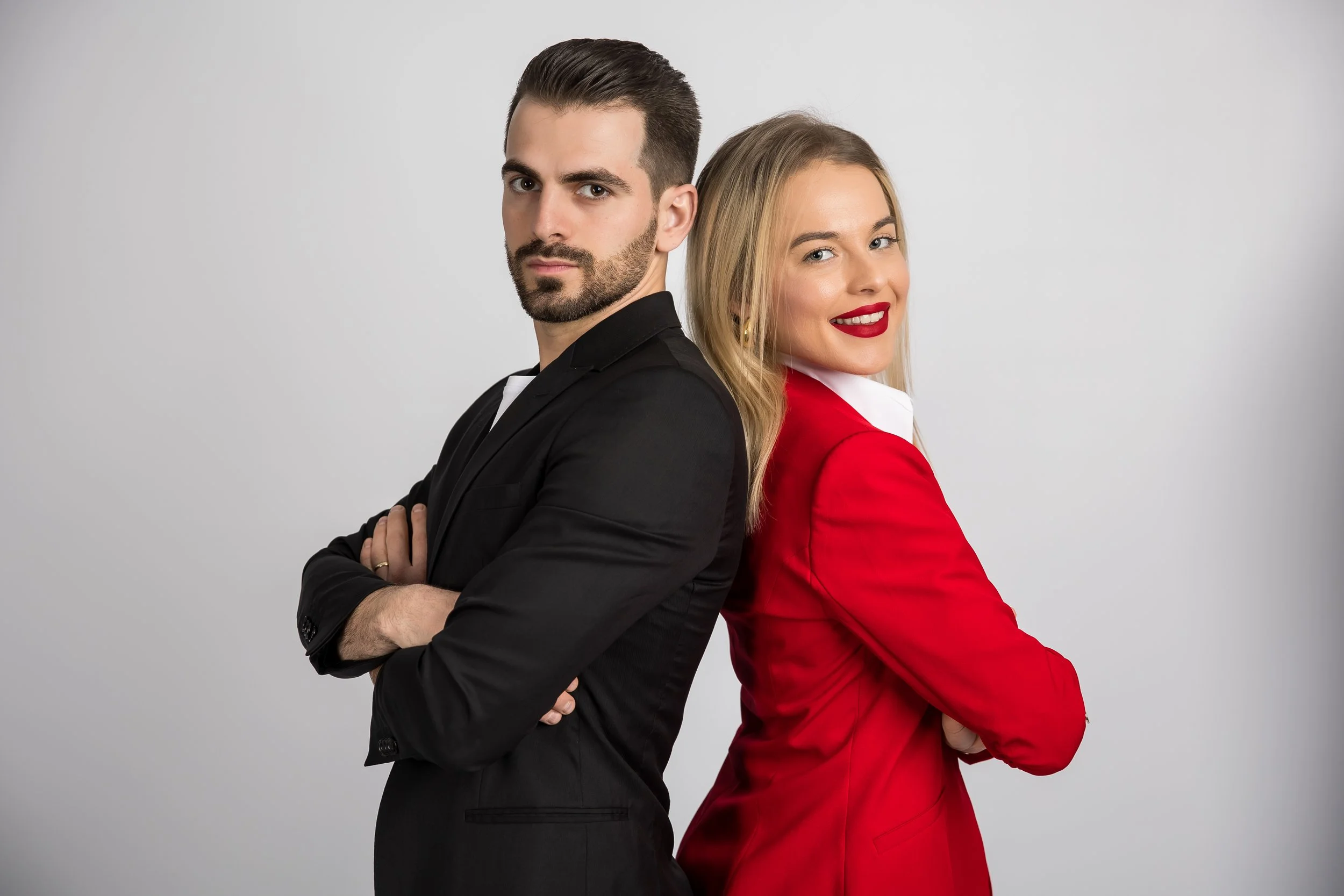 A man and a woman stand back-to-back with arms crossed, the man wearing a black suit and the woman wearing a red blazer, both looking at the camera against a plain light gray background.