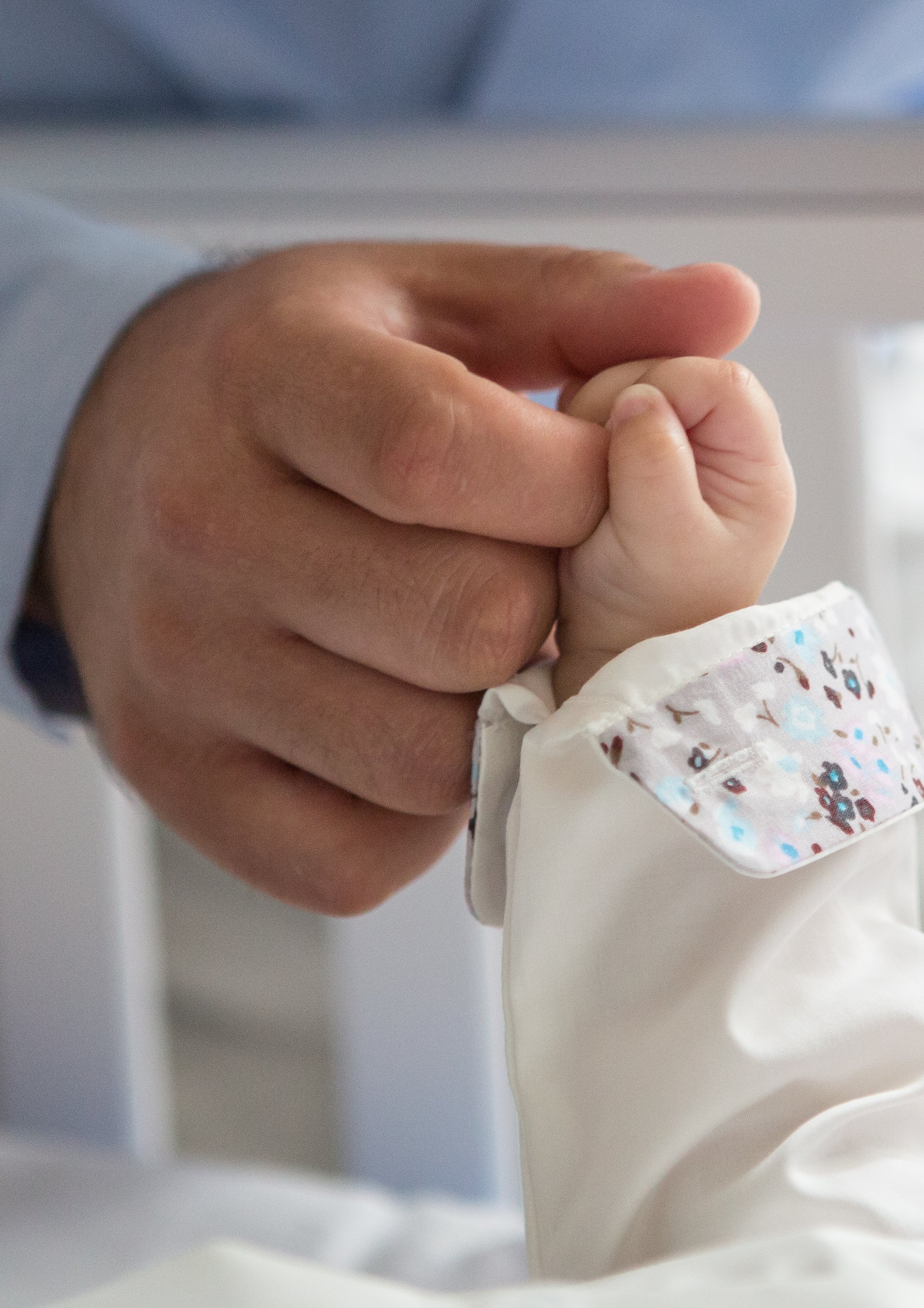 Close-up of a parent holding a newborn baby's tiny hand. The baby is wearing a white garment with colorful patterns.