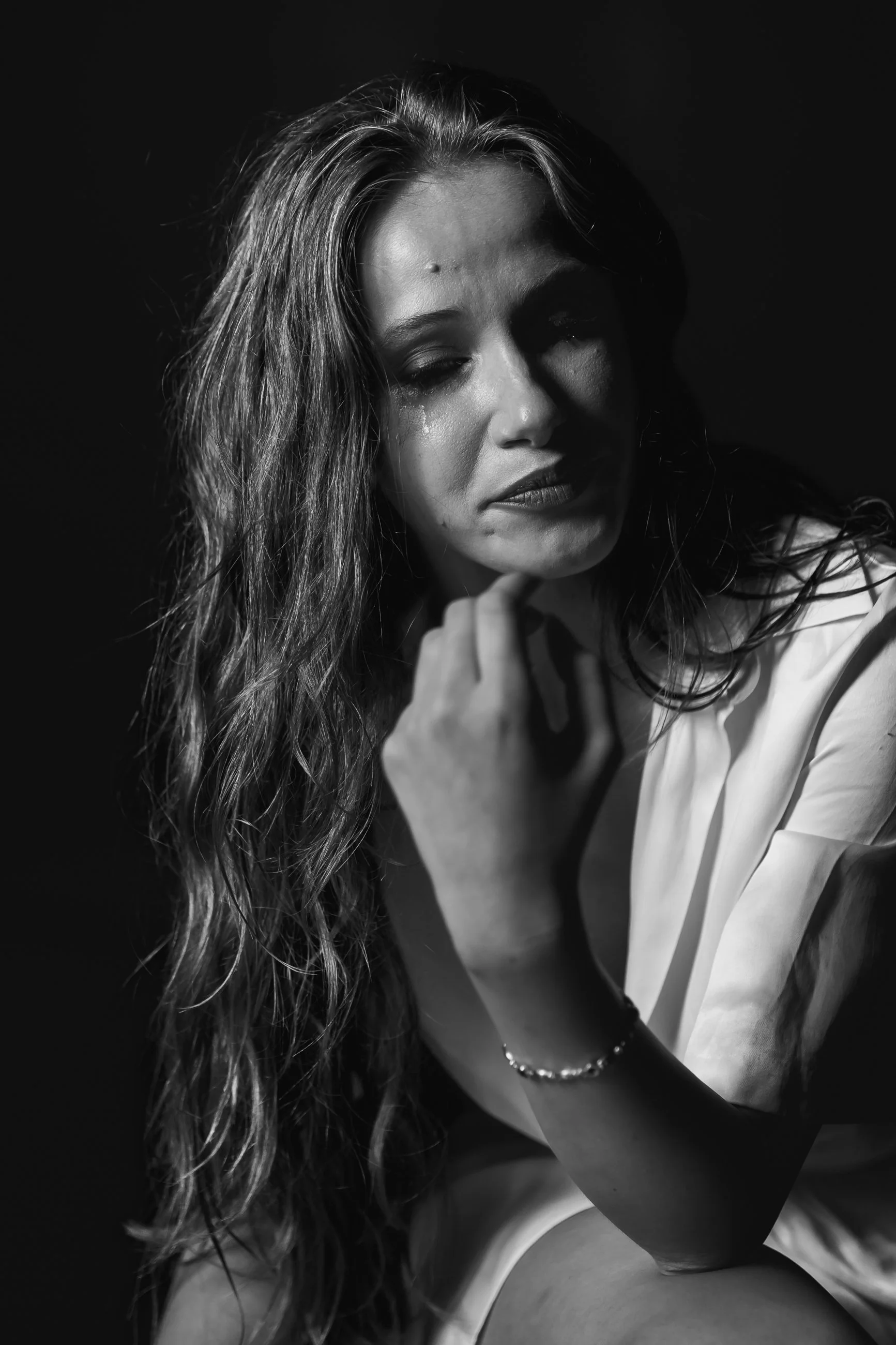 A black and white close-up photo of a woman with long, wavy hair, appearing emotional, with tears on her face, gazing down, wearing a white shirt, and resting her chin on her hand.