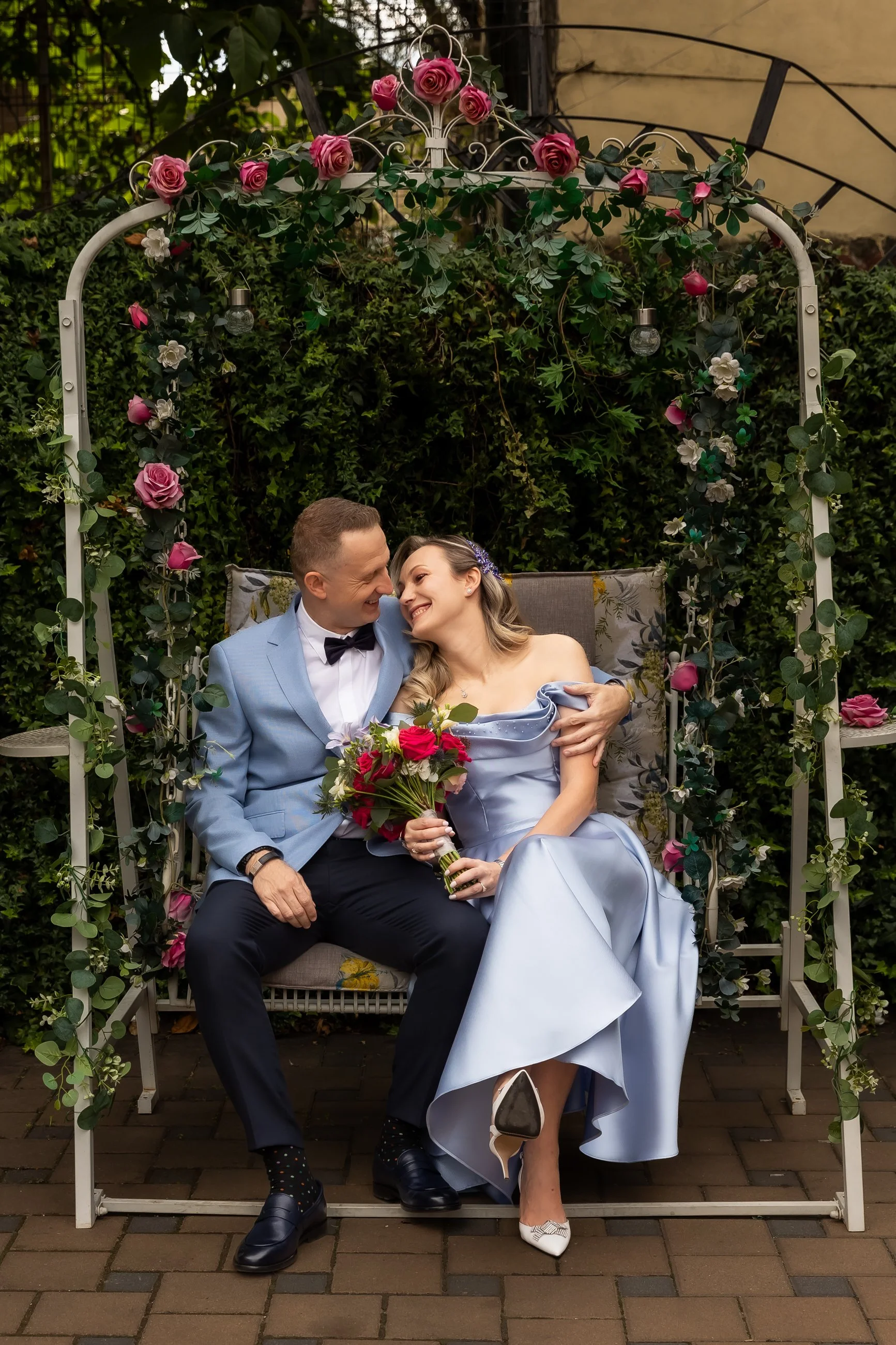 A joyful couple sitting close together on a decorated bench outdoors, surrounded by green foliage decorated with pink roses and white flowers, smiling and looking into each other's eyes, with the woman holding a bouquet of pink and white roses.