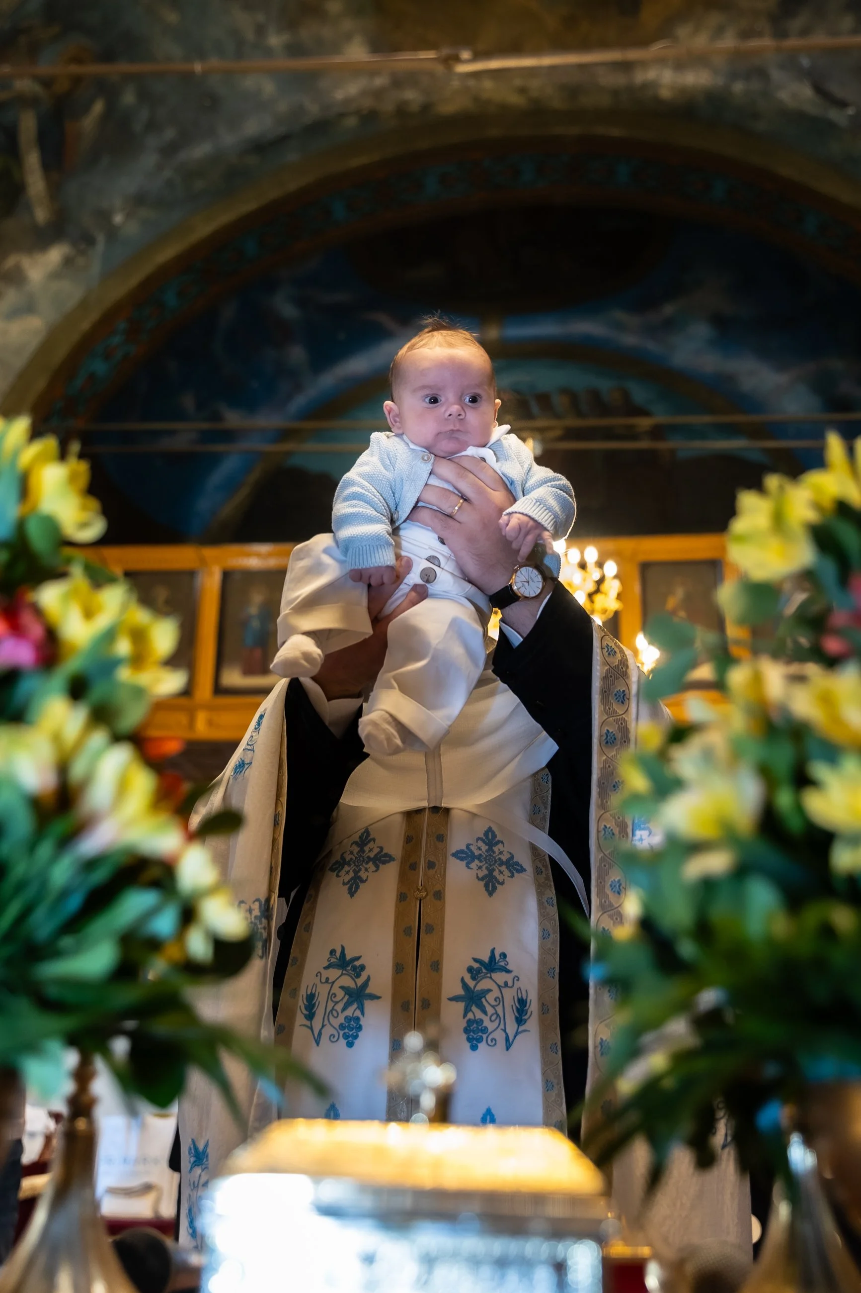 A baby being held up during a religious ceremony inside a church with floral decorations and an ornate ceiling in the background.