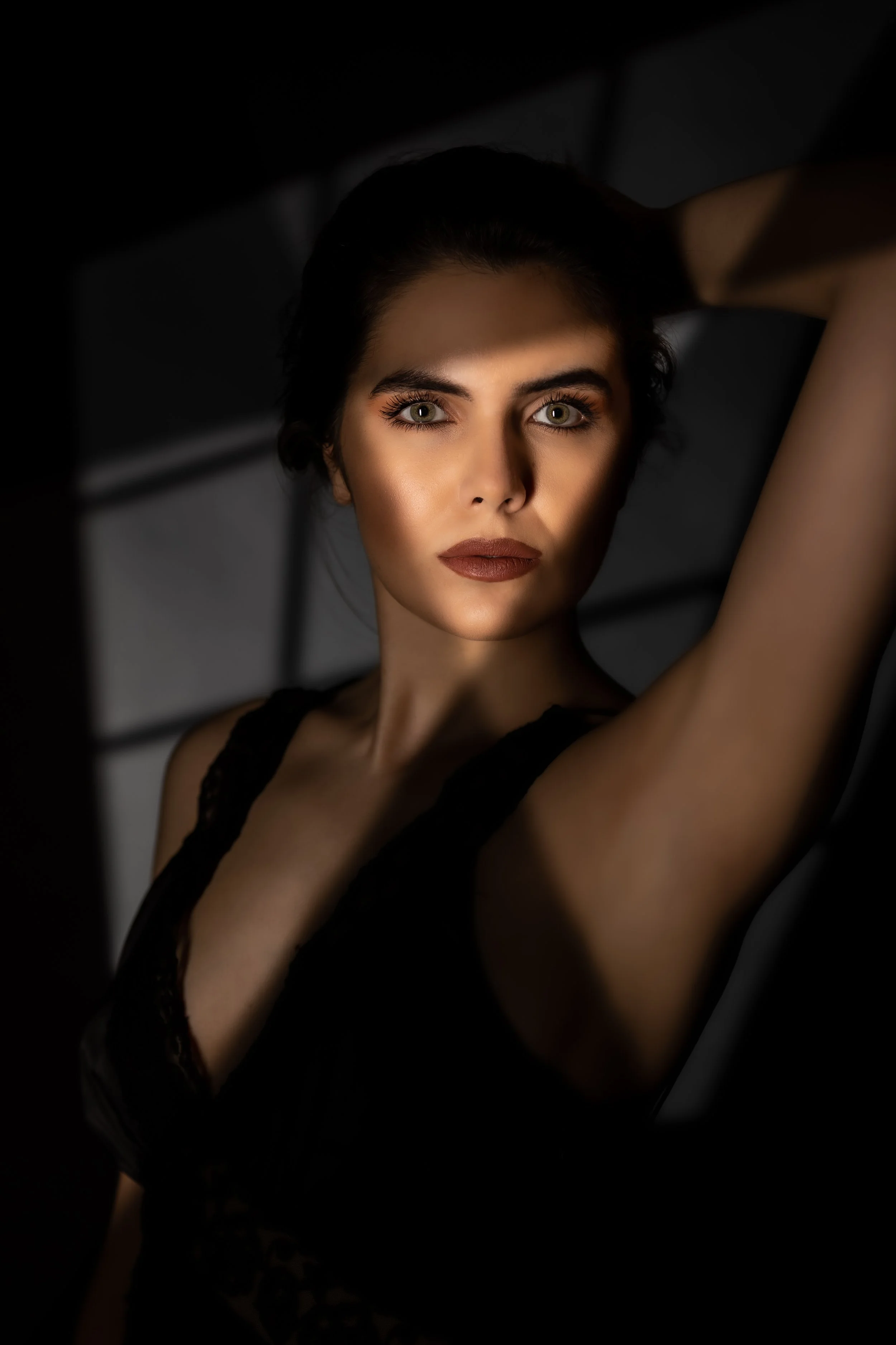 A woman with fair skin and striking green eyes, wearing makeup with bold lipstick, posing with her arm raised behind her head in a dark setting.