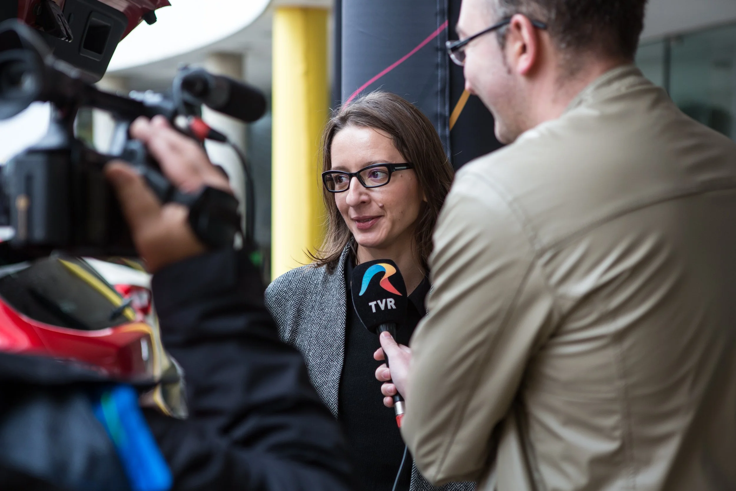 A woman with glasses being interviewed by a man holding a TVR microphone, with camera equipment in the foreground.
