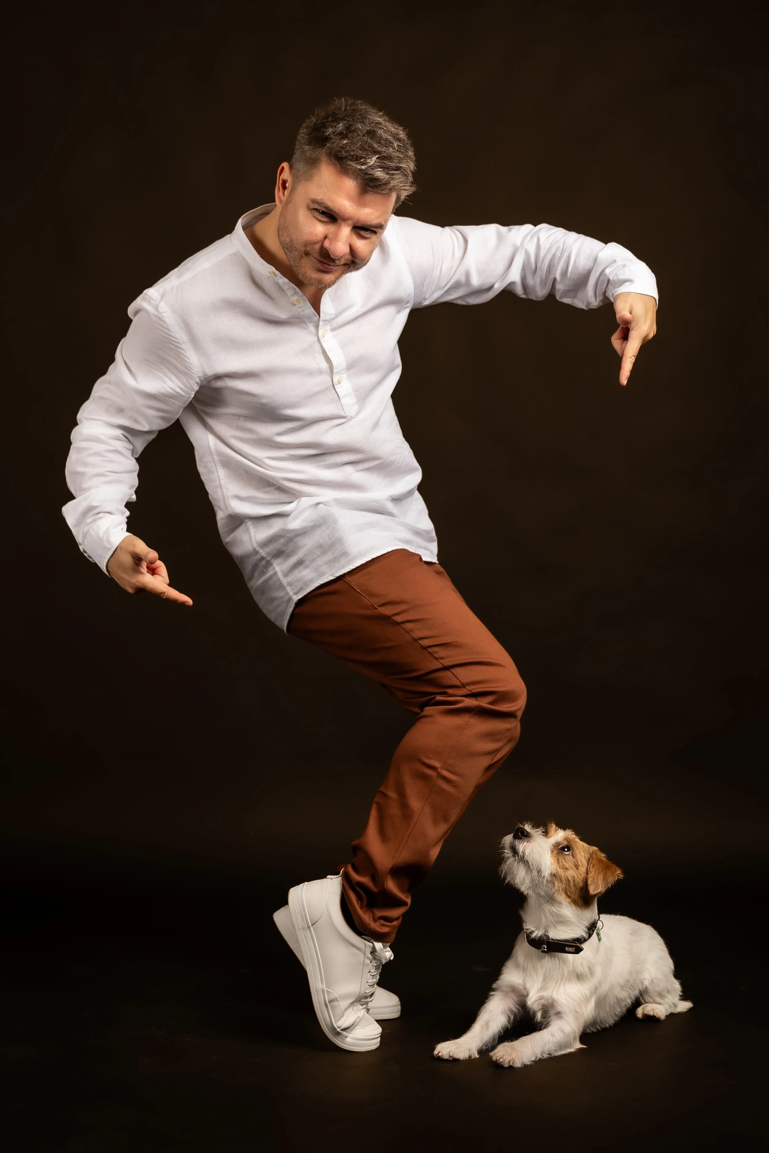A man with grey hair, wearing a white button-up shirt, brown pants, and white sneakers, pointing down at a puppy with his right hand, while the puppy is sitting on the floor looking up at him, against a dark background.