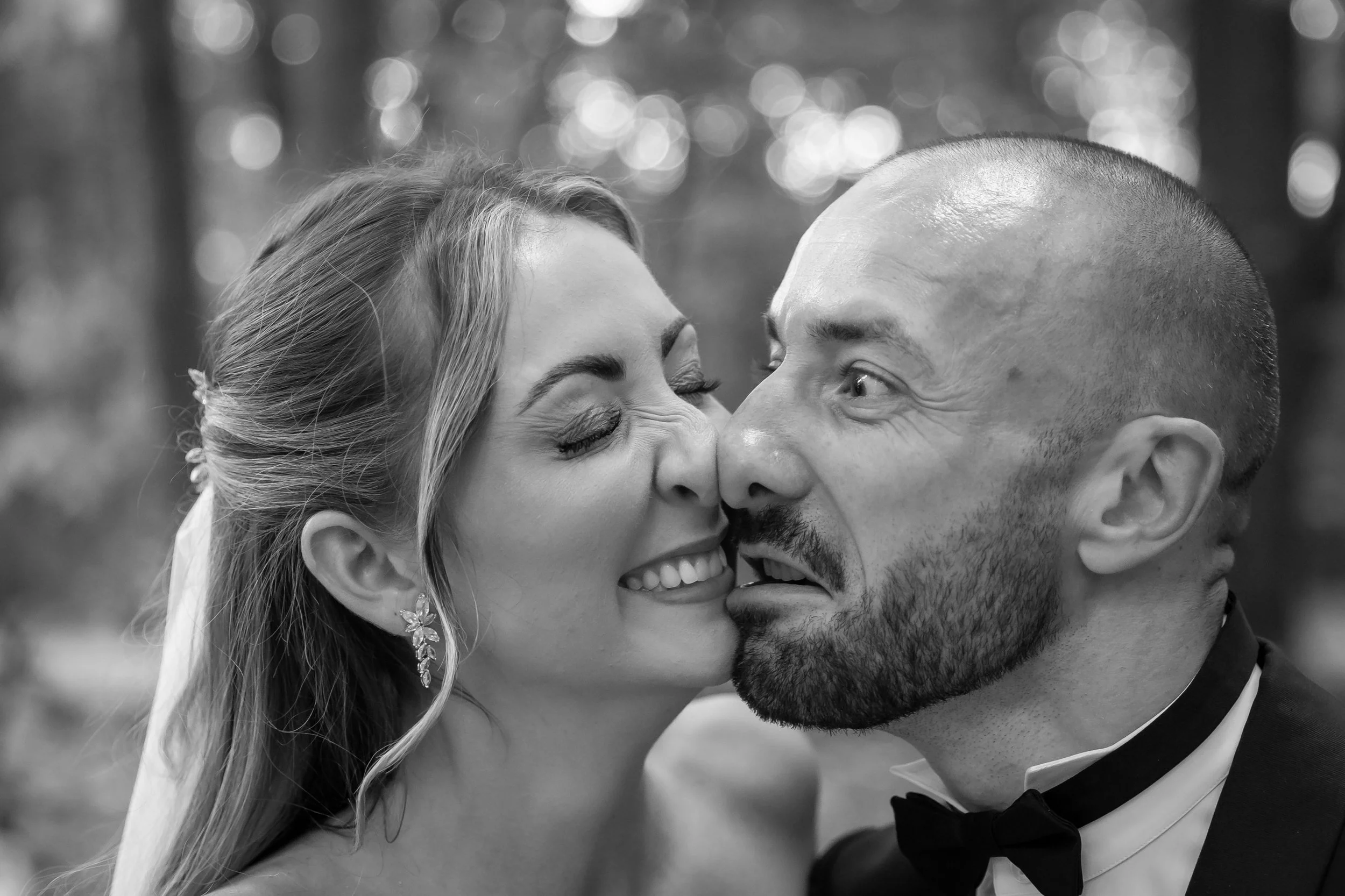 A close-up black and white photo of a couple smiling and touching noses with eyes closed, outdoors with blurred lights in the background.